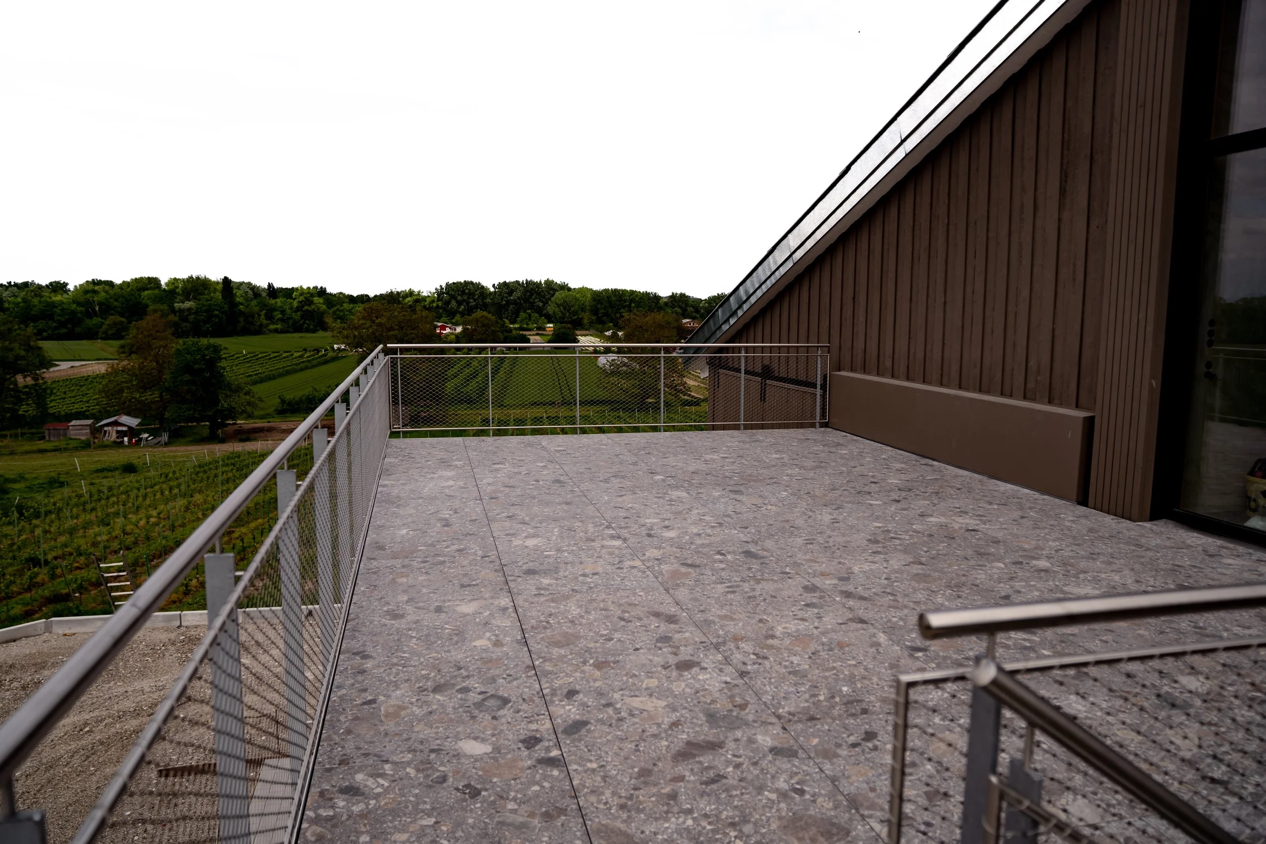 Balkon mit Steinboden und Blick auf grüne Felder und Bäume im Hintergrund, an der Seite eine Hauswand aus Holzplatten.