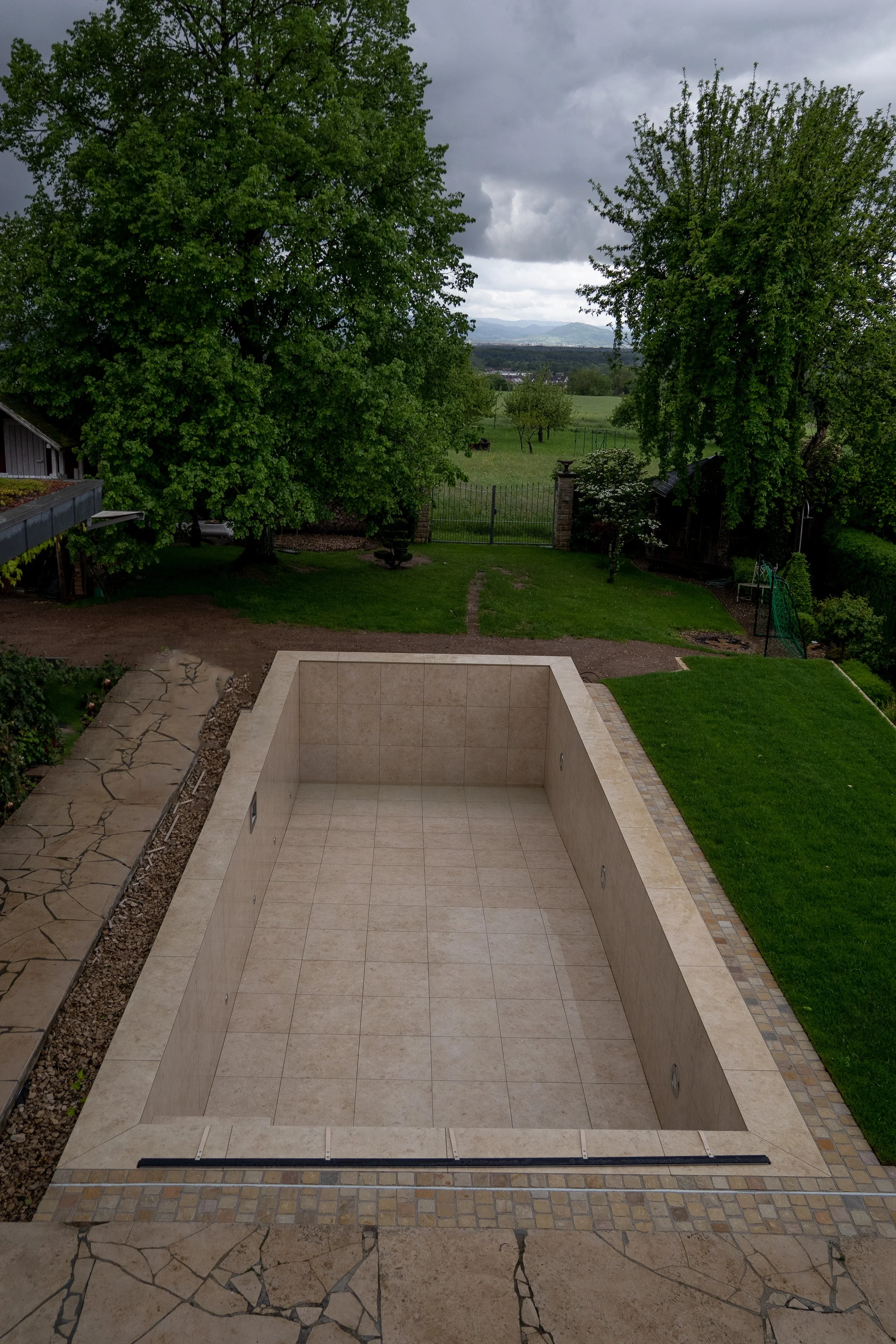 Leeres, privates Swimmingpool im Garten, umgeben von Kies und Fliesen, mit grünem Rasen und Bäumen im Hintergrund bei bewölktem Himmel.