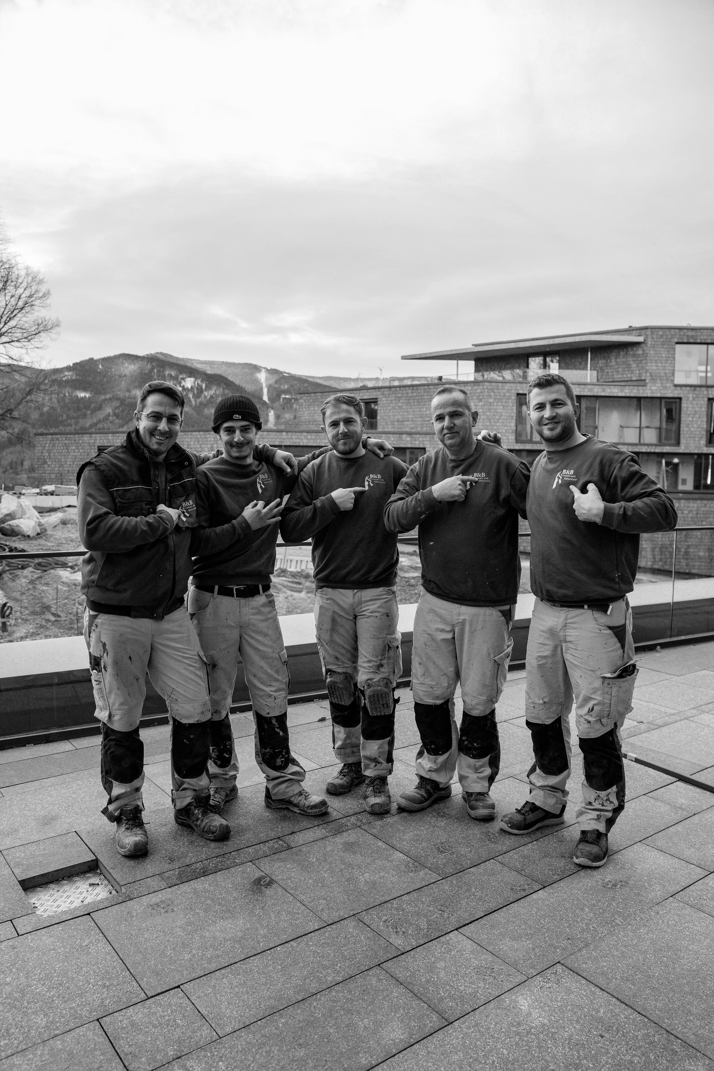 Fünf Handwerker stehen auf einer Terrasse vor einer modernen Wohnung im Freien, lachen und zeigen auf ihre Arbeit, mit Berglandschaft im Hintergrund, schwarz-weiß Aufnahme.