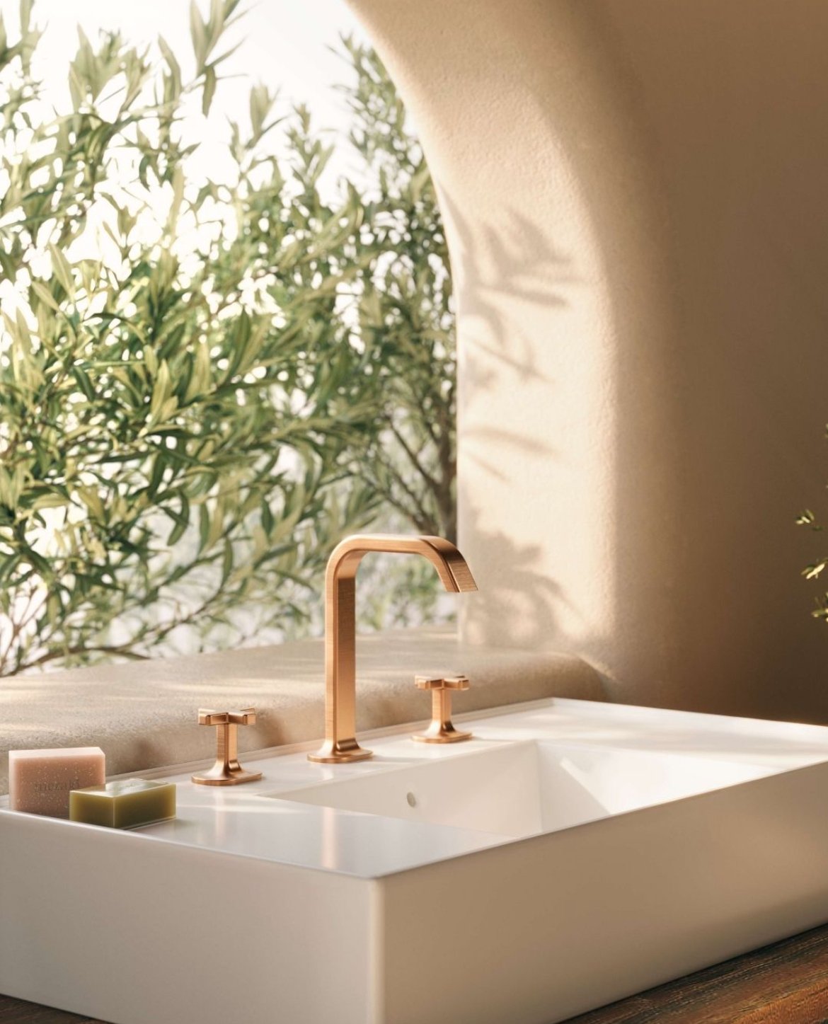 Elegante Badezimmerwaschbecken mit rötlich-goldener Armatur, rosa und grüne Seifen auf Holzablage, mit Blick auf Olivenbaum im Hintergrund.