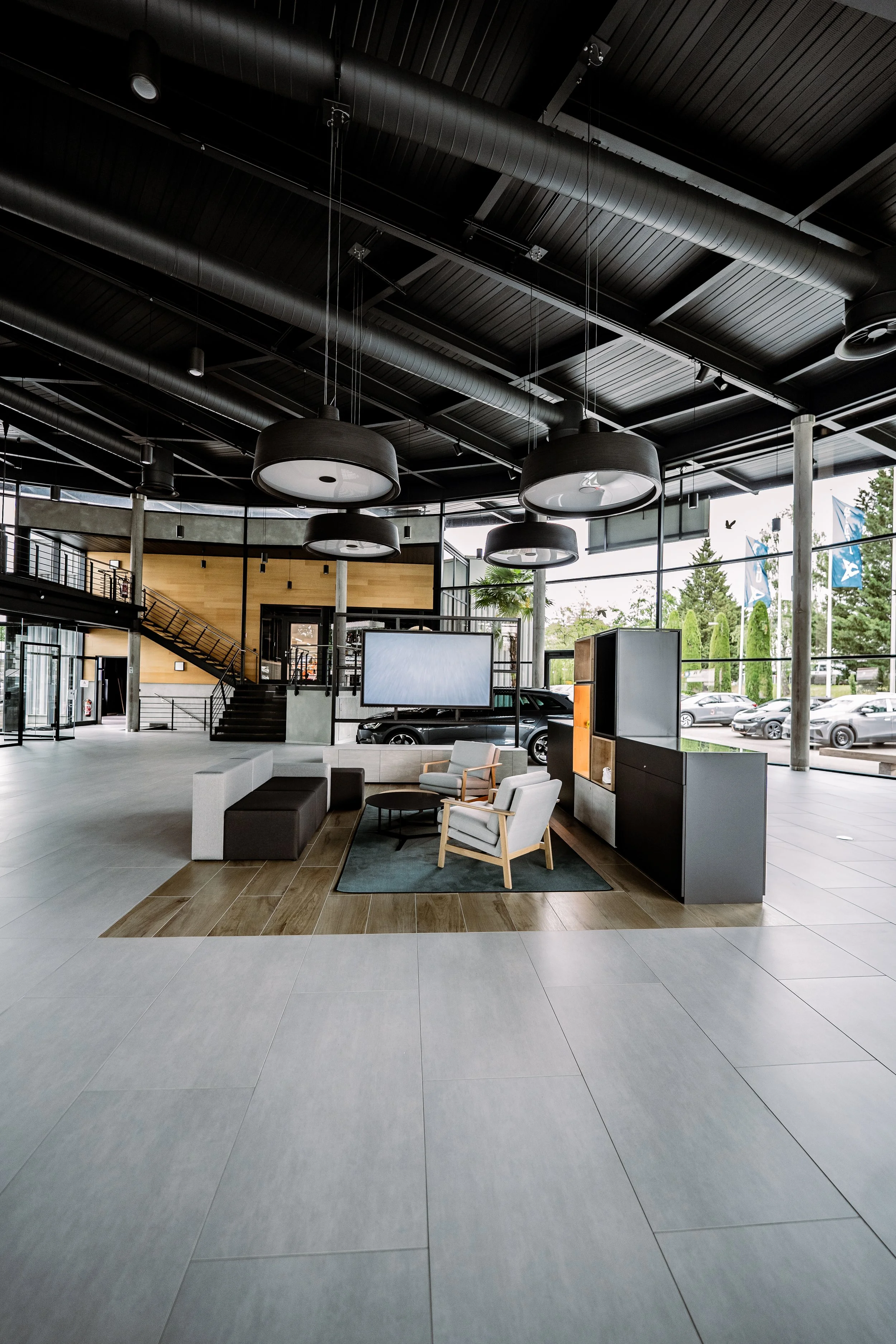 Modernes, offenes Autohaus mit Sitzbereich, großen Fenstern, schwarzen Deckenleuchten, Parkplätzen im Hintergrund.
