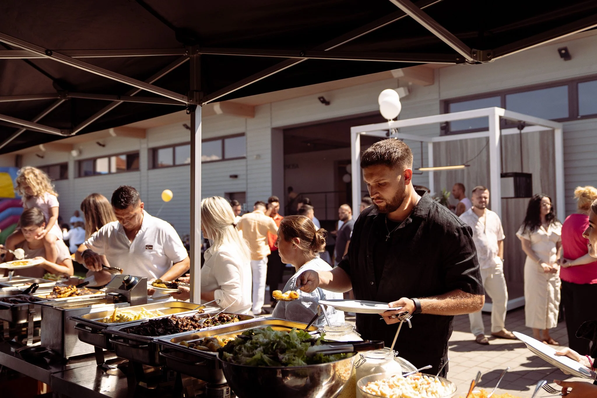 Menschen beim Buffet auf einer Gartenparty, mit Essen und Getränken, im Freien vor einem modernen Gebäude.