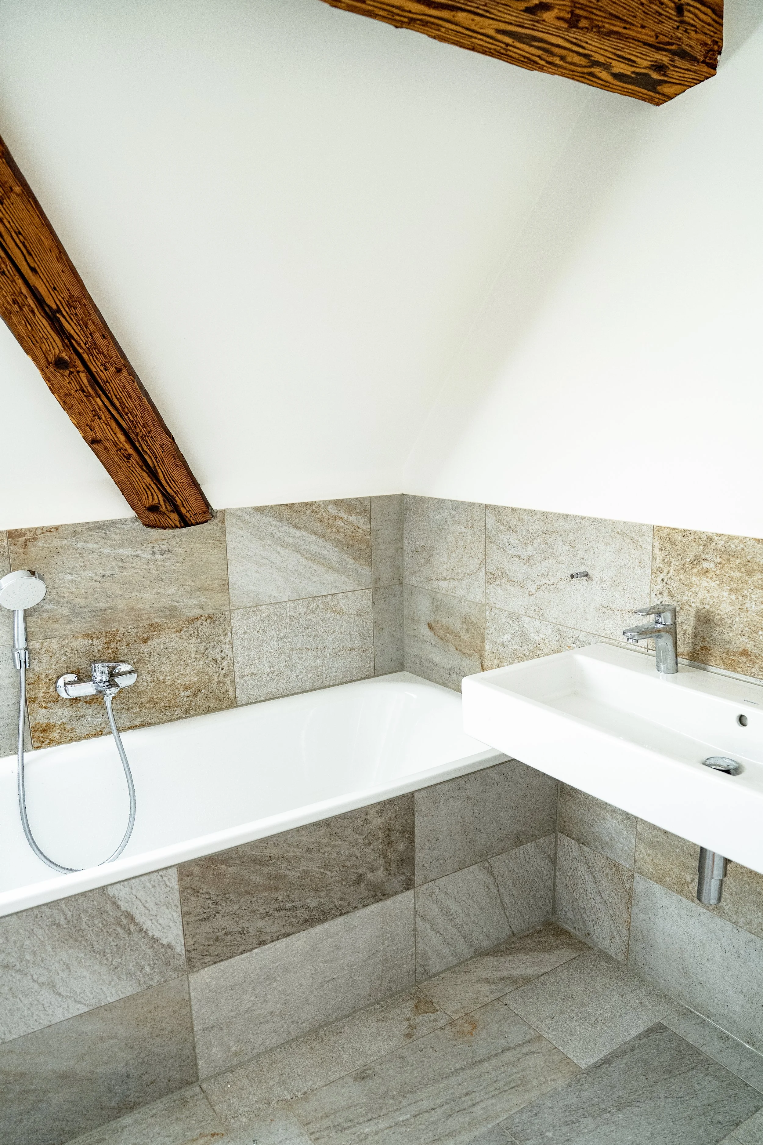 Badezimmer mit Badewanne und Waschbecken, Wände und Boden aus grauen Steinfliesen, Dachbalken aus Holz, weiße Wände, modernes Design.