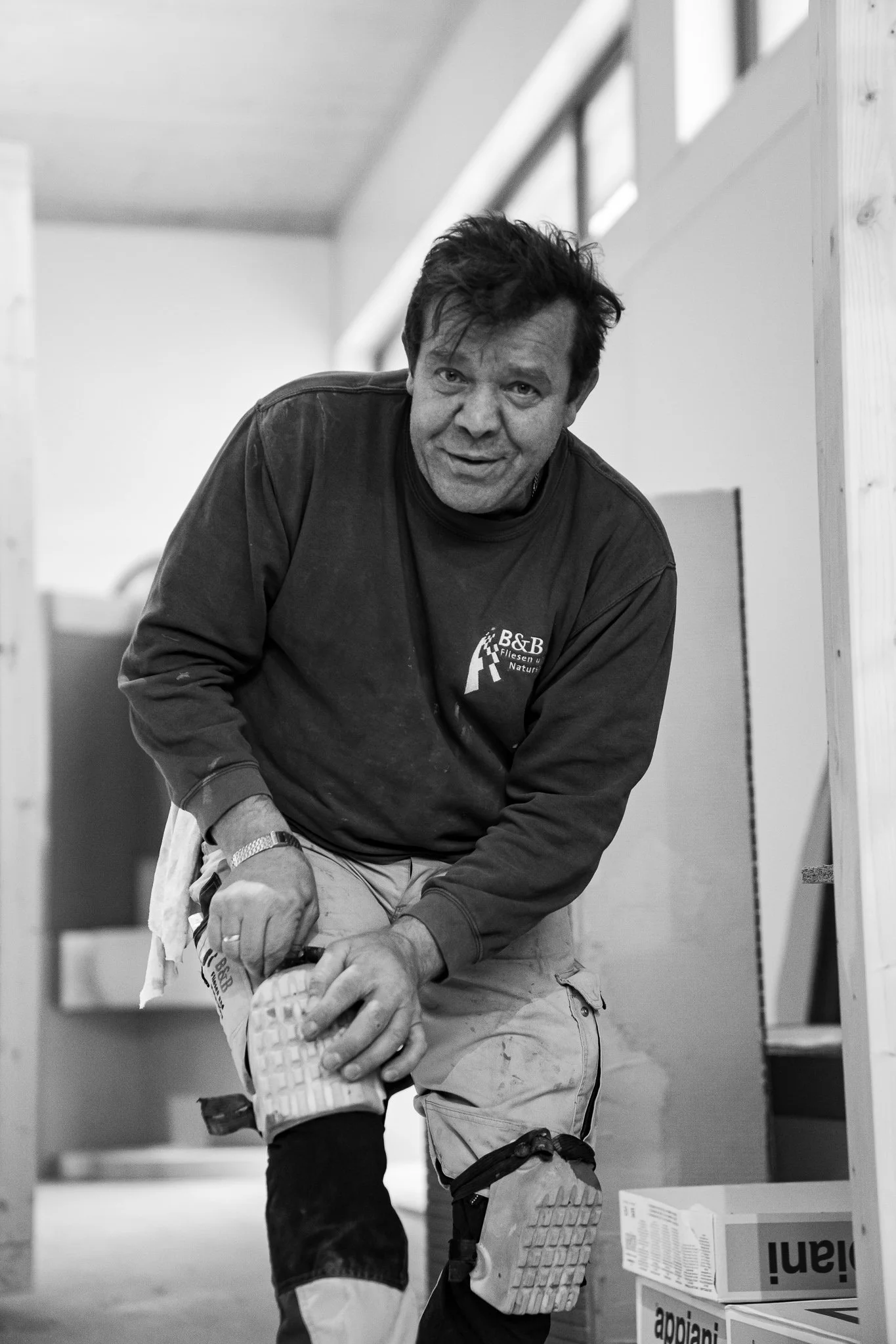 Ein Mann kniet auf einem Baugerüst, trägt Arbeitskleidung und schaut in die Kamera.