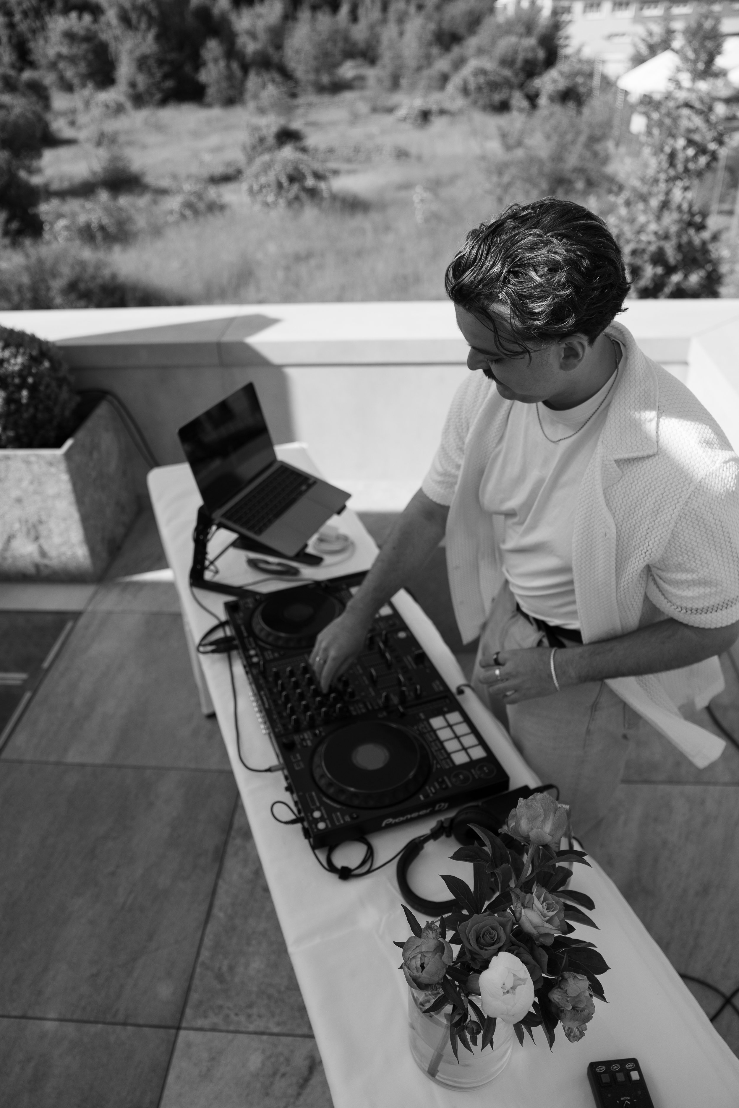 Person beim Auflegen von Musik mit DJ-Equipment auf einem Balkon, im Vordergrund eine Vase mit Blumen, im Hintergrund grüner Garten