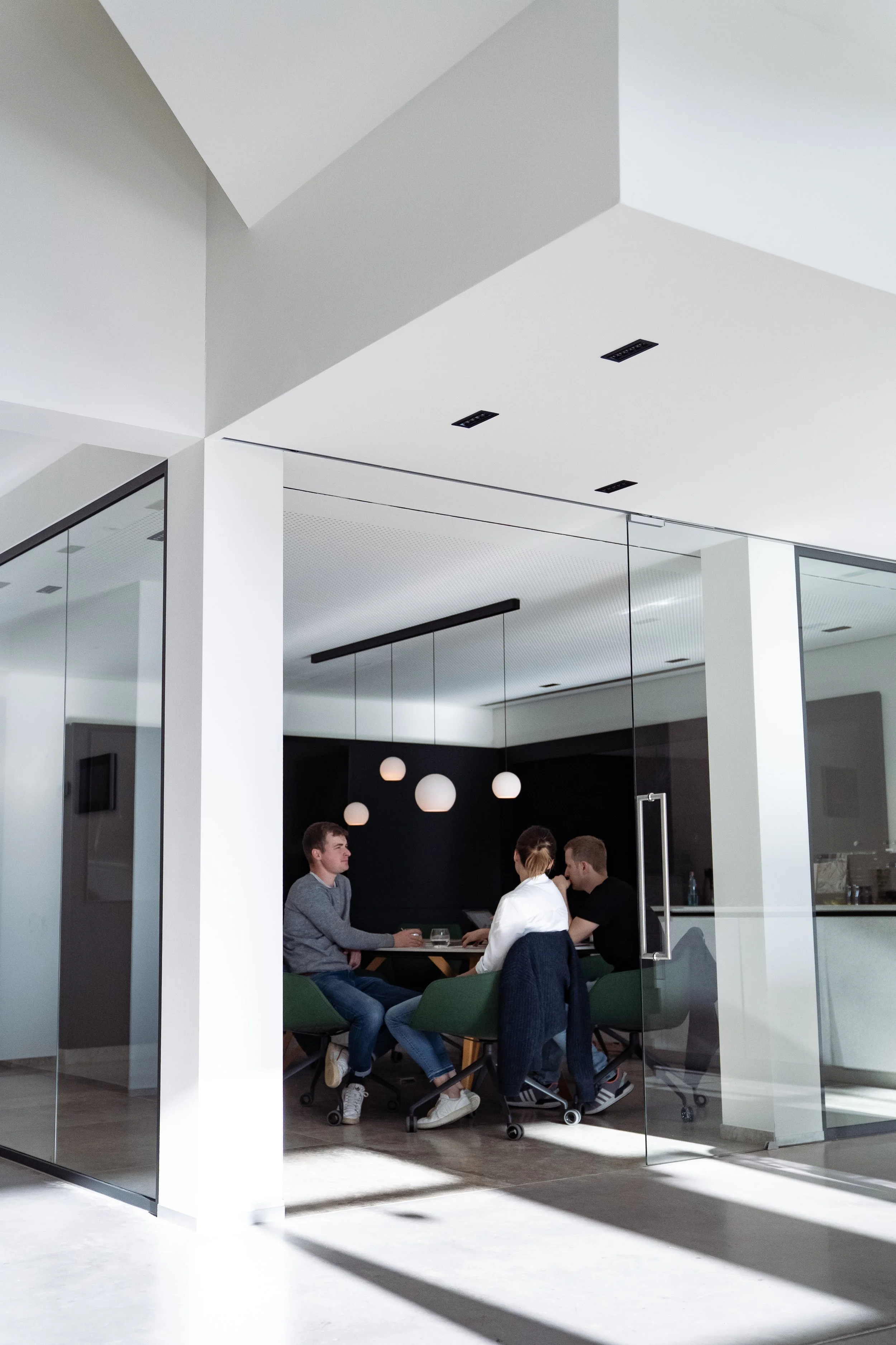 Drei Menschen sitzen an einem Tisch in einem modern gestalteten Raum mit schwarzen Wänden, weißen Kugellampen und Glaswänden.