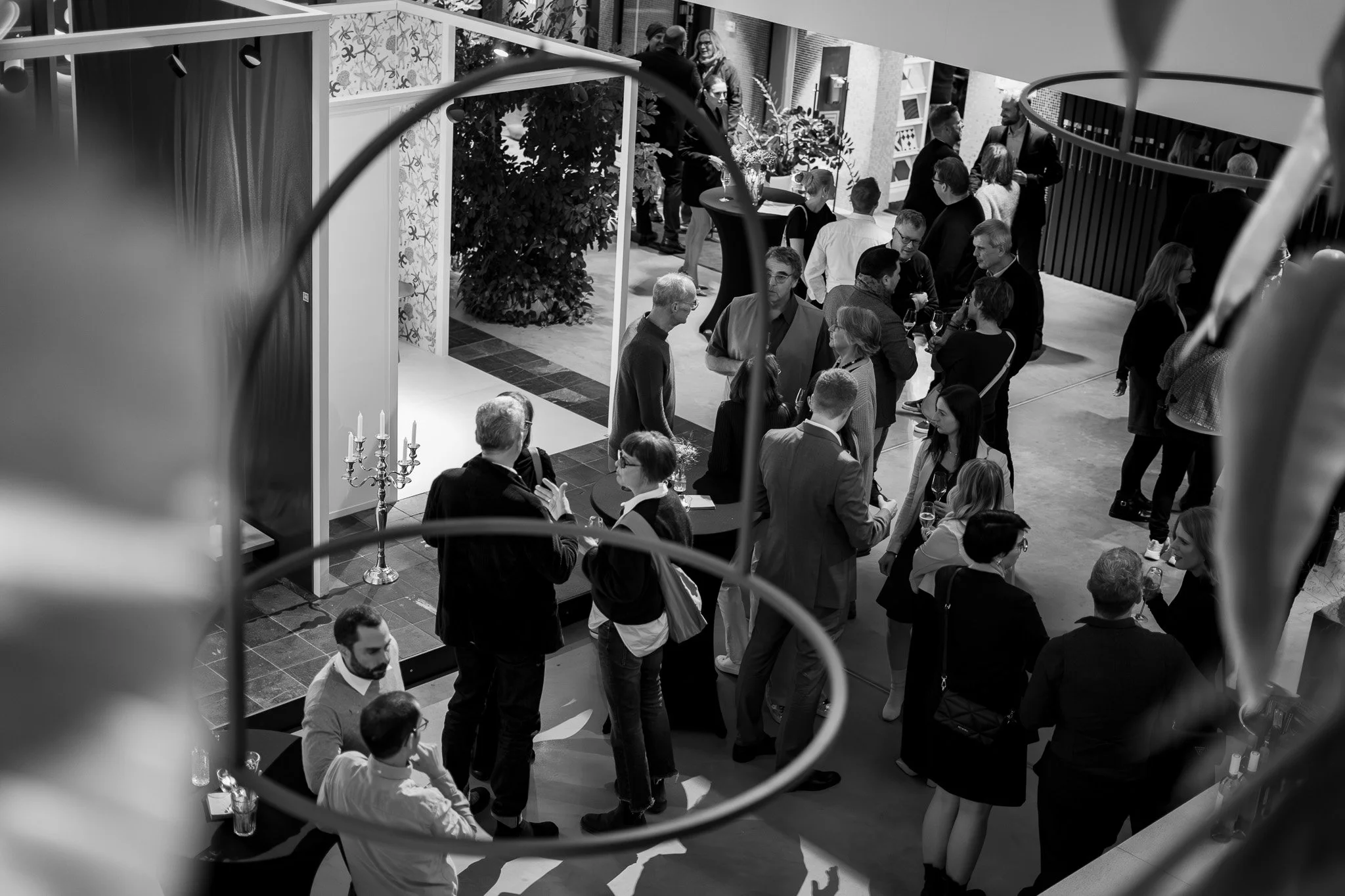 Menschen versammeln sich auf einer Veranstaltung im Innenraum, einige unterhalten sich, während andere Getränke halten, mit Dekorationen wie Kerzenleuchtern und Pflanzen im Hintergrund.