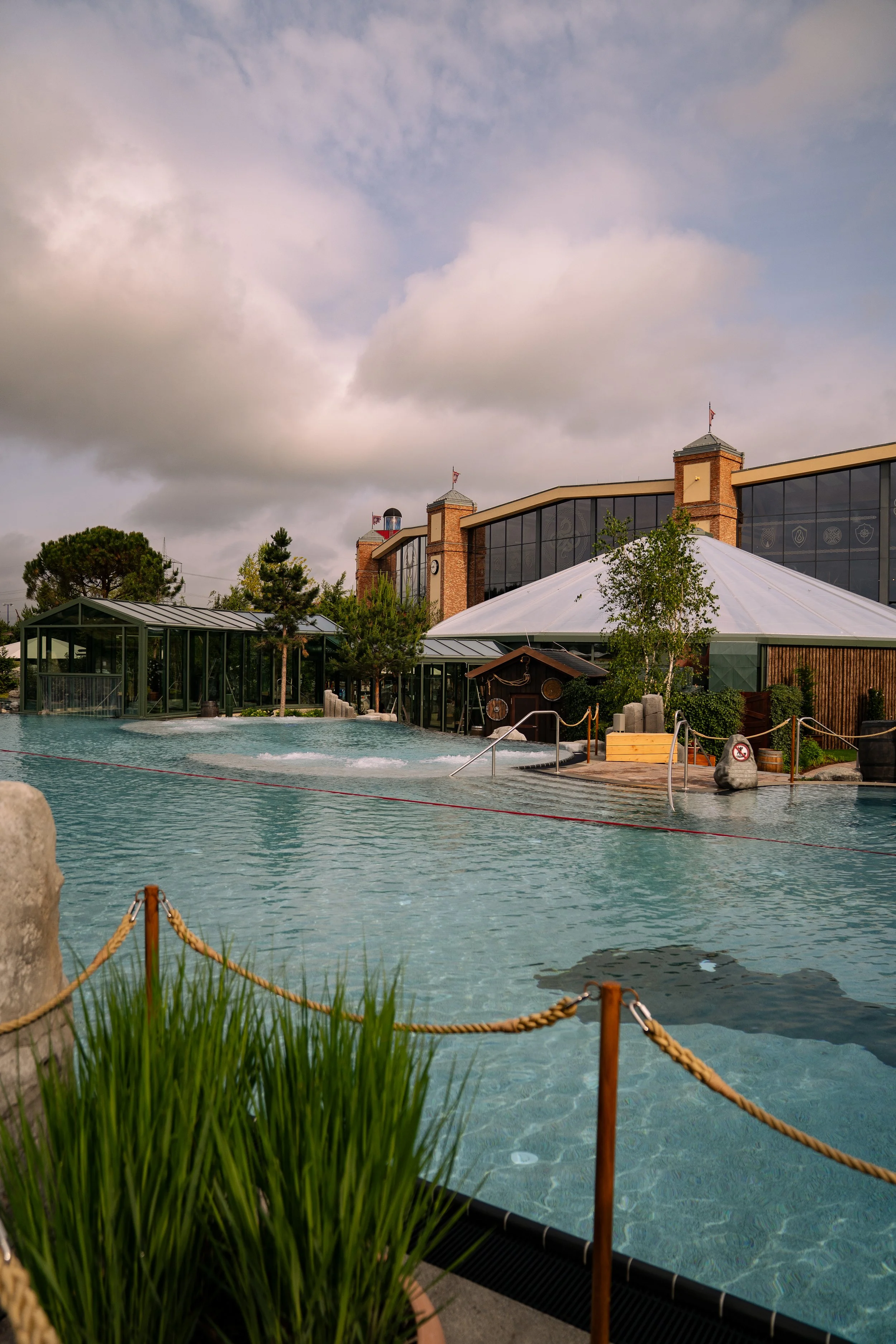 Ein Außenbereich eines Spas oder Wellnesscenters mit einem beheizten Pool, umgeben von Glasarchitektur und grüner Vegetation bei bewölktem Himmel.