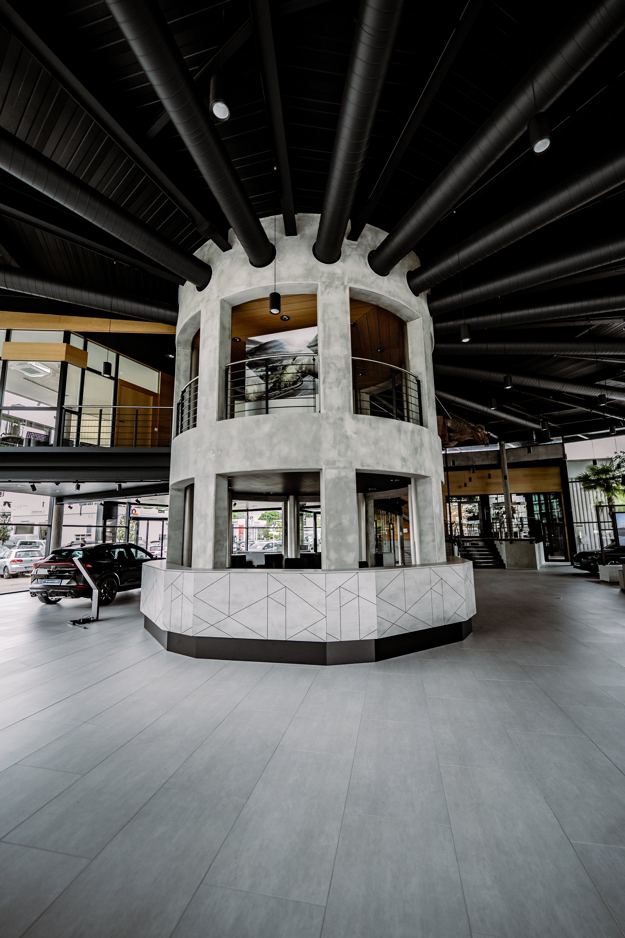 Innenraum eines modernen Autohaus mit einem zentralen runden Empfangstresen, schwarzen Deckenrohren und großen Glasfenstern.