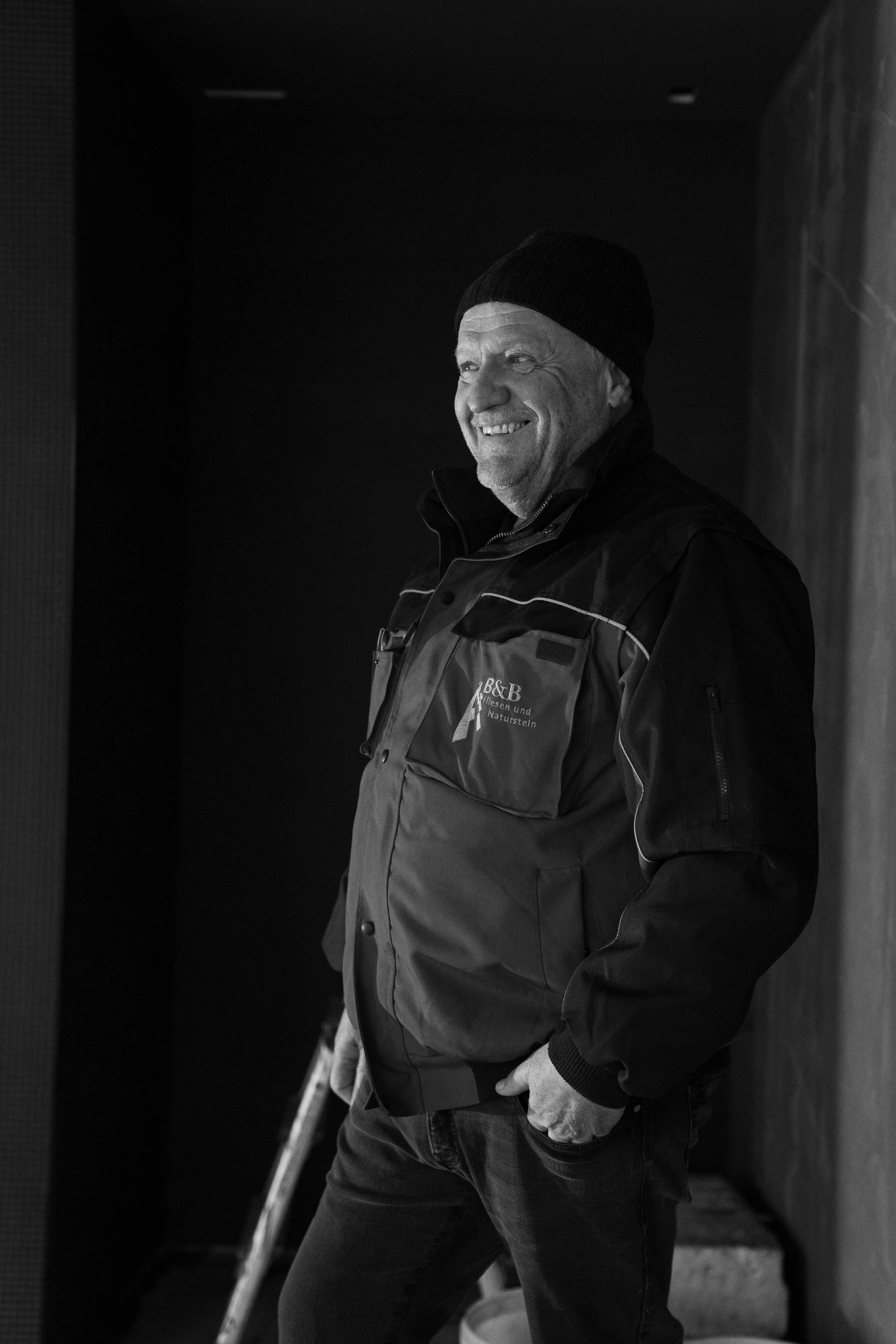 Ein älterer Mann lächelt und trägt eine Mütze, schwarze Jacke und Jeans, steht in einem dunklen Raum an einer Wand.