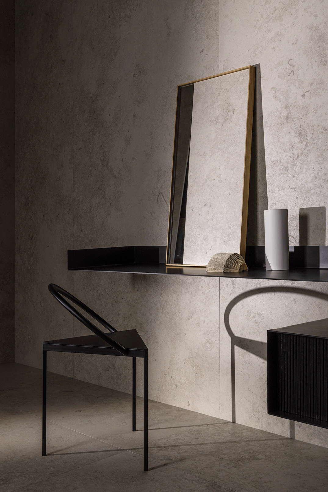 Ein dunkler Tisch mit einem großen, hochkant stehenden Spiegel, einer weißen Vase, einem kleinen Buch und einem schwarzen Box. Ein schwarzer Stuhl steht davor. Die Szene ist minimalistisch gestaltet, mit einem Betonwand-Hintergrund und weichem Licht.