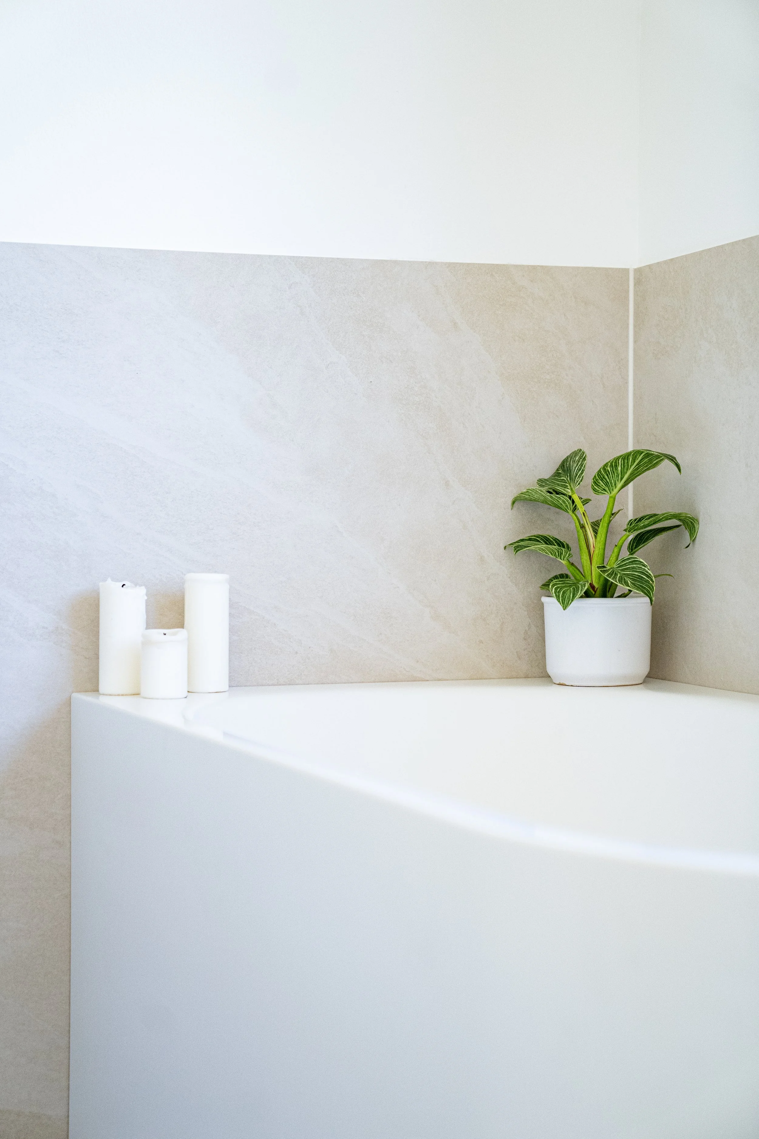 Ein weißer Badewannenrand mit drei weißen Kerzen und einer Grünpflanze in einem weißen Top in einer Ecke mit beigefarbenen Fliesen.