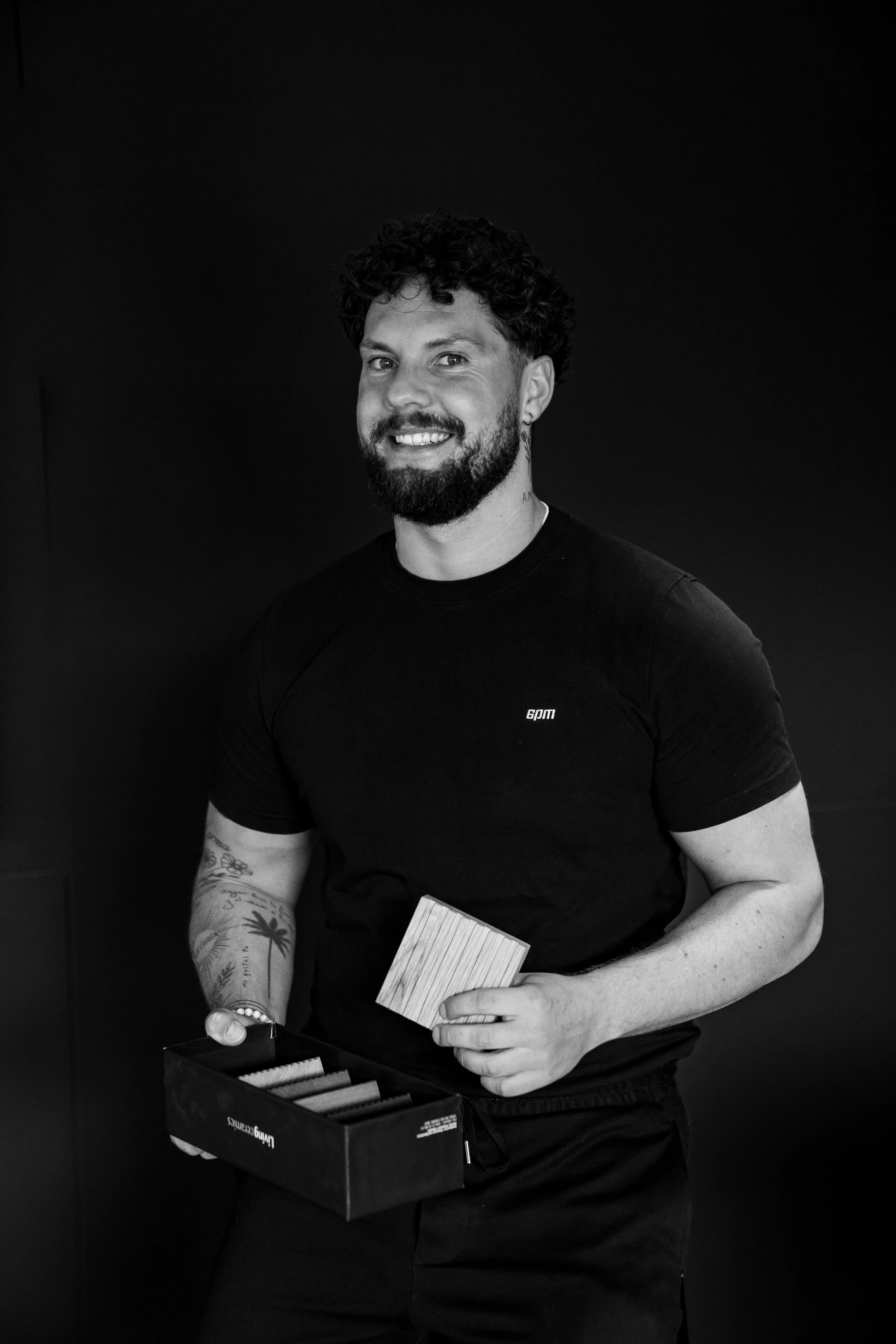 Lächelnder Mann mit Bart und Tattoos, hält Holzstück und steht vor schwarzem Hintergrund.