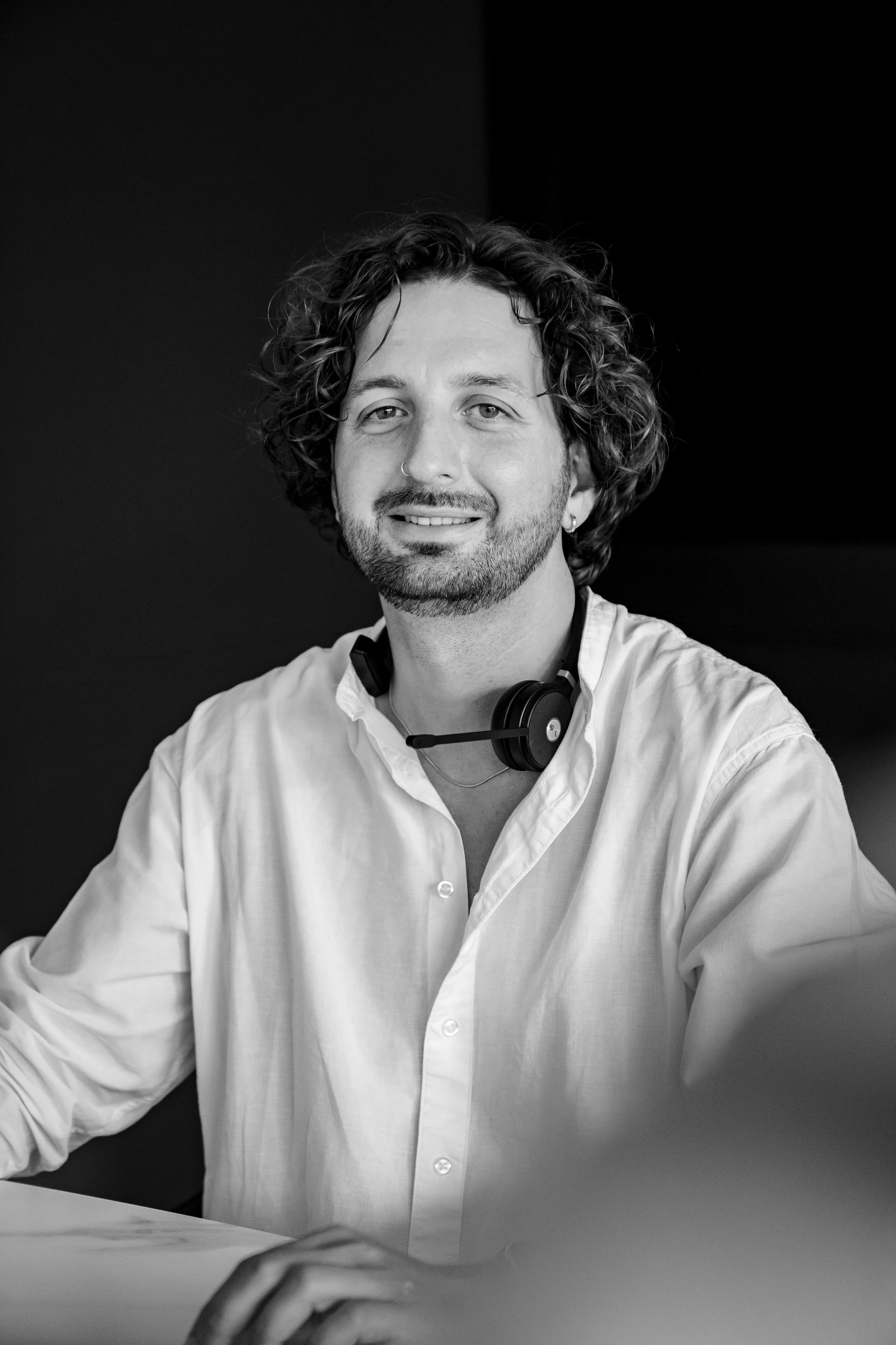 Mann mit lockigem Haar, im weißen Hemd, trägt ein Headset, lächelnd, im schwarzen und weißen Foto.