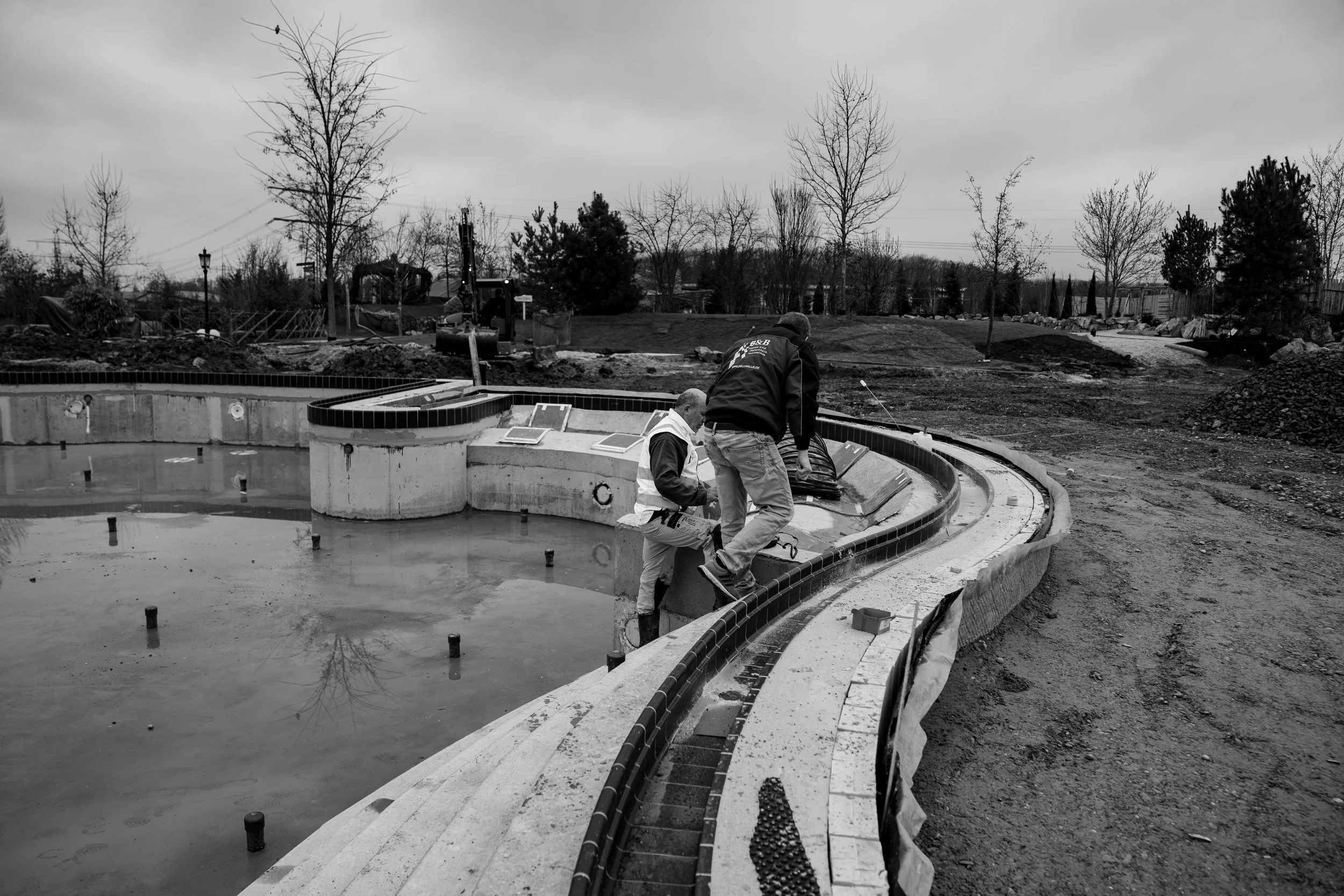 Zwei Bauarbeiter installieren eine Wasserbecken-Fußleiste auf einer Baustelle im Freien bei bewölktem Himmel.