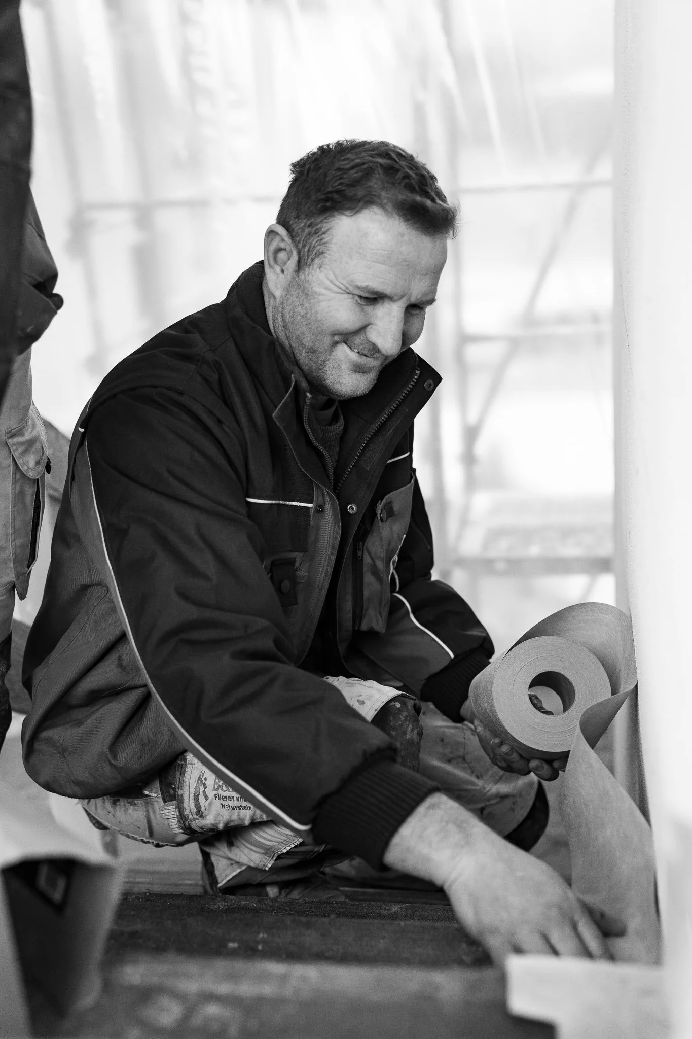 Mann mit dunkler Jacke, der auf dem Boden sitzt und Baumaterial, wahrscheinlich eine Isolierung, hält.