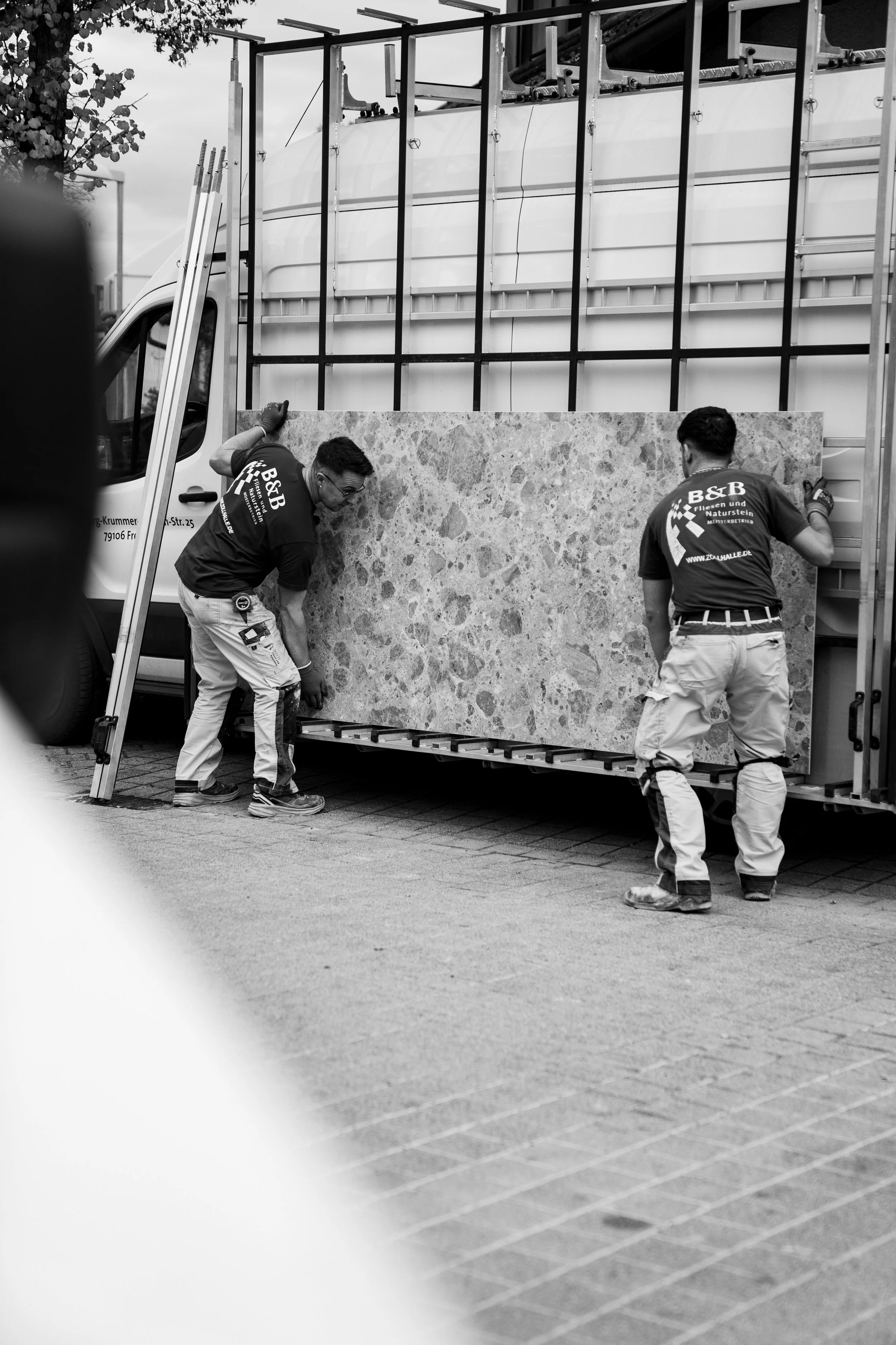 Zwei Männer heben eine große Steintafel auf einen Zeilenwagen, vor einem Lieferwagen.