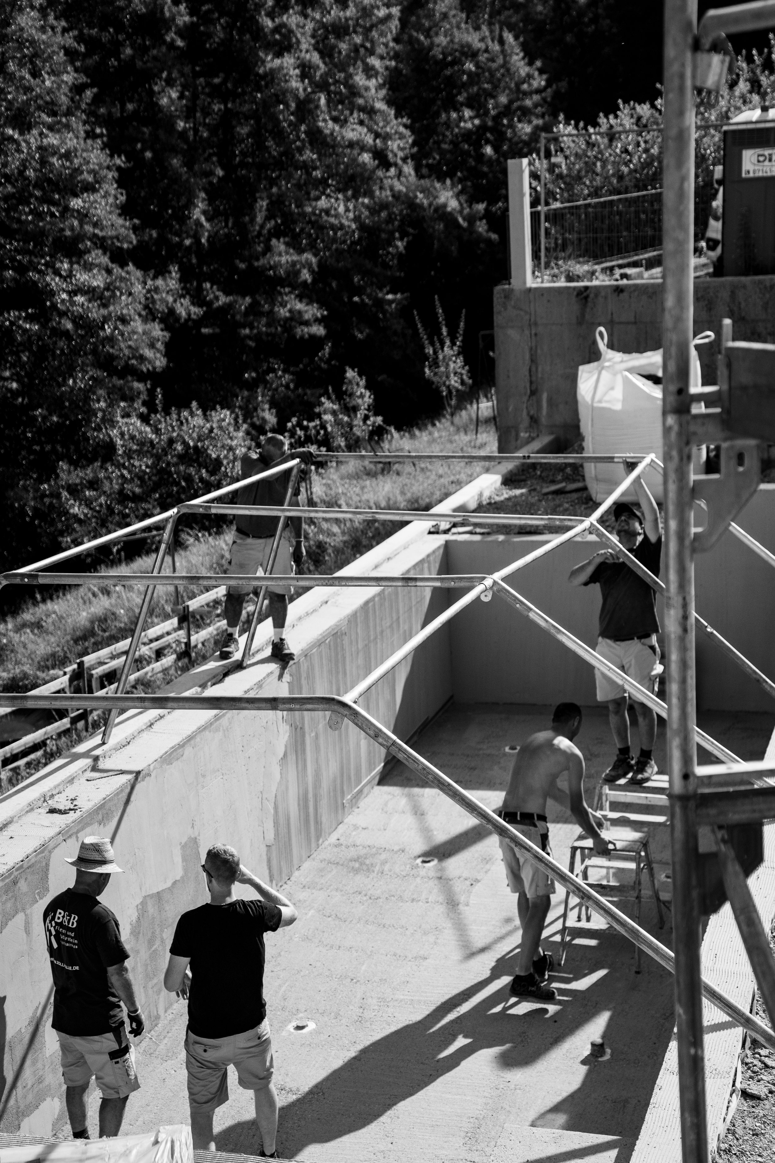 Menschen bauen eine Pool- oder Beckenwand aus Beton auf einer Baustelle, im Hintergrund Bäume, einige im Gespräch, andere bei der Arbeit