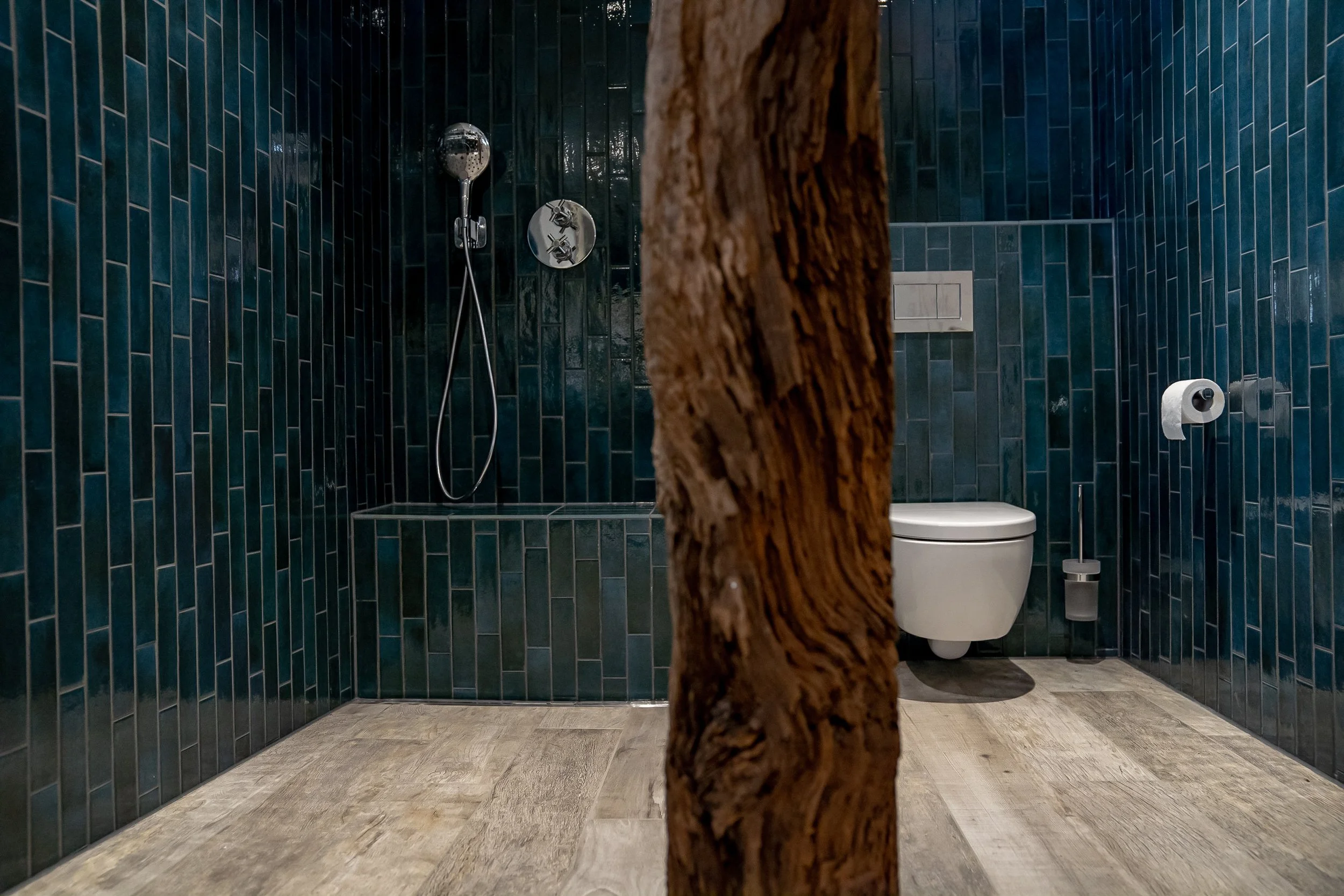 Moderne Badezimmer mit dunklen Wandfliesen, Dusche und wandhängendem WC, getrennt durch eine Baumstamm-Partition.