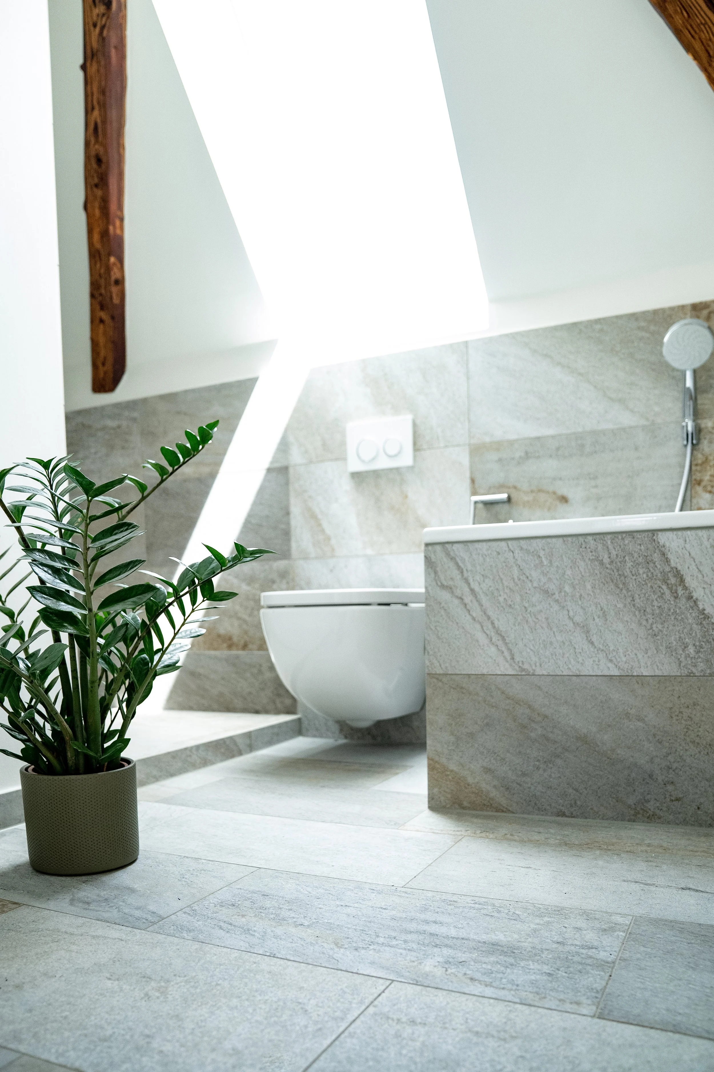 Ein modernes Badezimmer mit einem WC, einer Pflanze in einem Topf, Fliesenwänden und -böden, einer Dusche und einem Holzträger an der Decke.