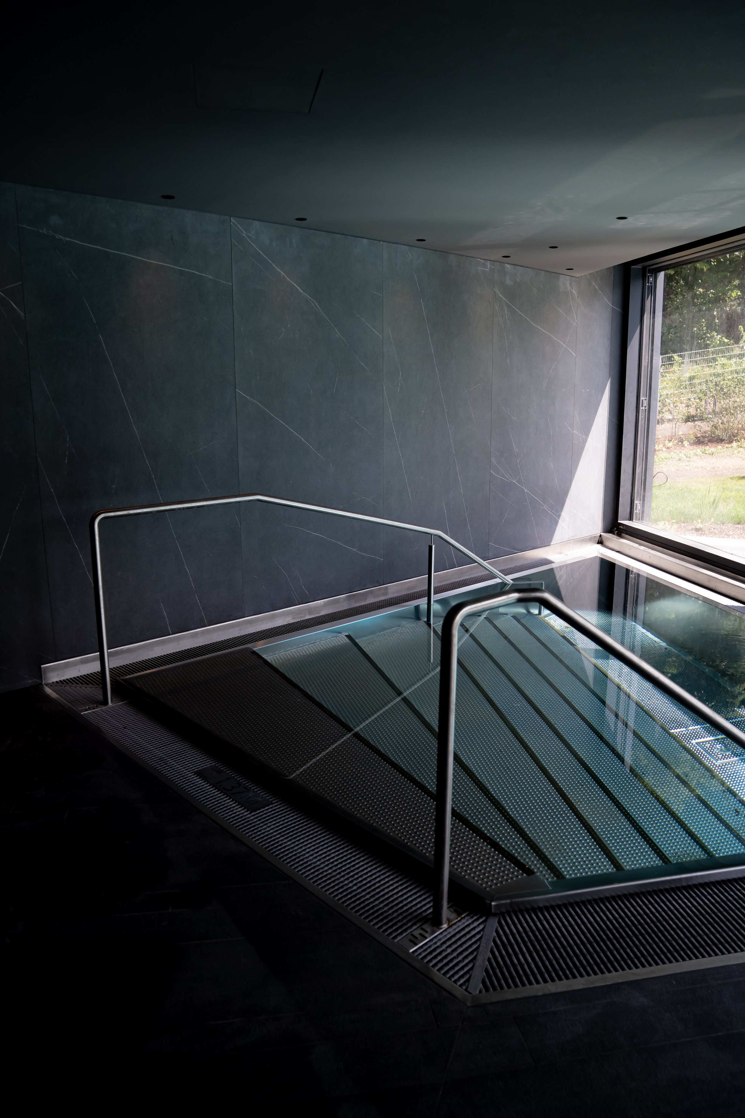 Innenansicht eines modernen Spa- oder Wellnessbereichs mit einem kleinen Pool, schwarzem Fliesenboden und dunkelgrauer Wand mit Marmorierung, großer Fensterscheibe, durch die Tageslicht fällt.