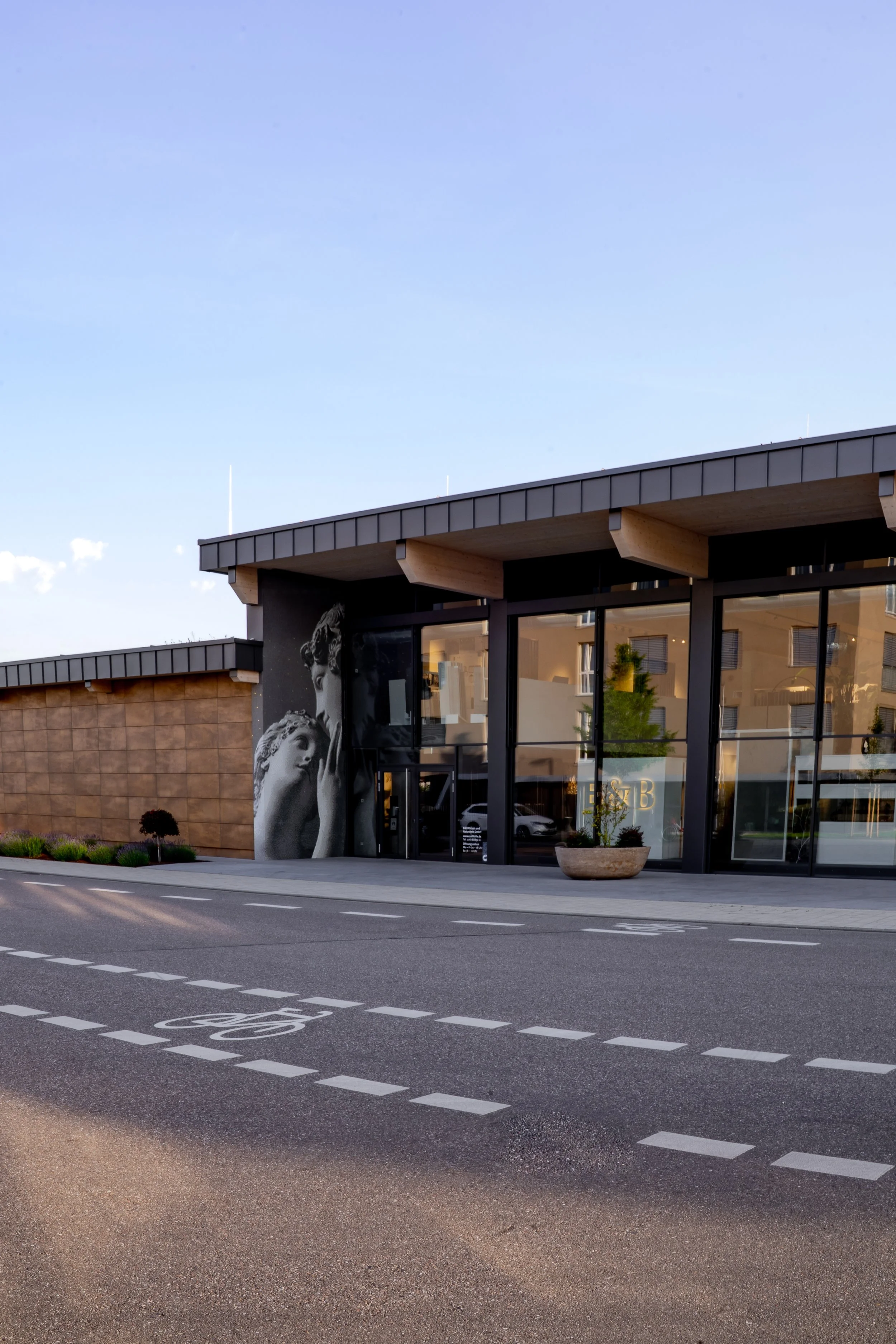 Moderne Glasfront eines Gebäudes mit Wandkunst eines Skulpturenpaares, umgeben von einem Fahrradstreifen und einem tiefer gelegenen Bürgersteig, bei Tageslicht.