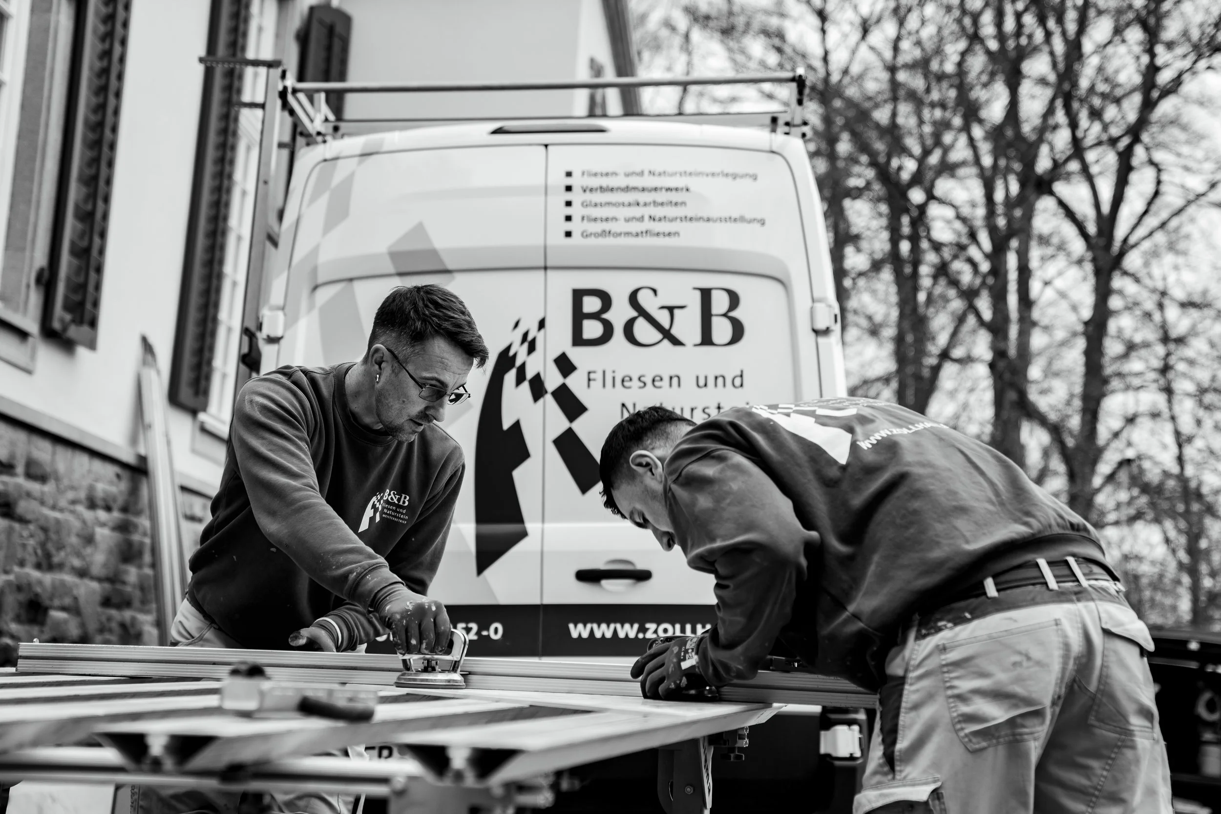 Zwei Männer arbeiten zusammen an einer Holzarbeit neben einem Firmenwagen im Freien.