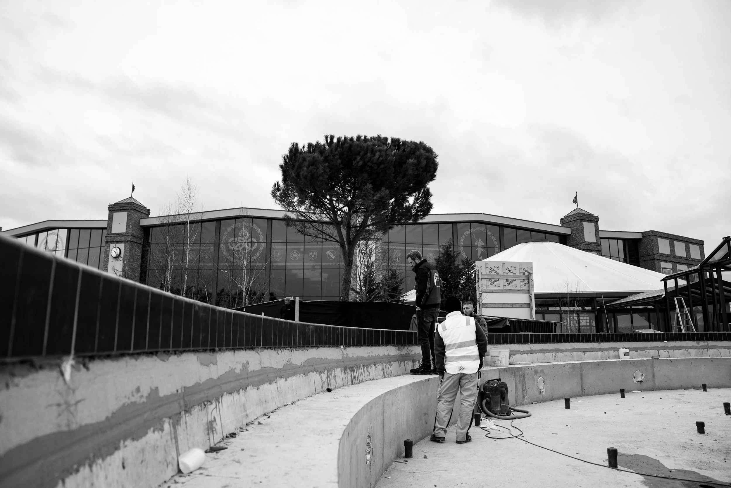Baustelle mit drei Arbeitern, einem elektrischen Werkzeug, einer weißen Kuppel und einem modernen Gebäude im Hintergrund in Schwarz-Weiß.