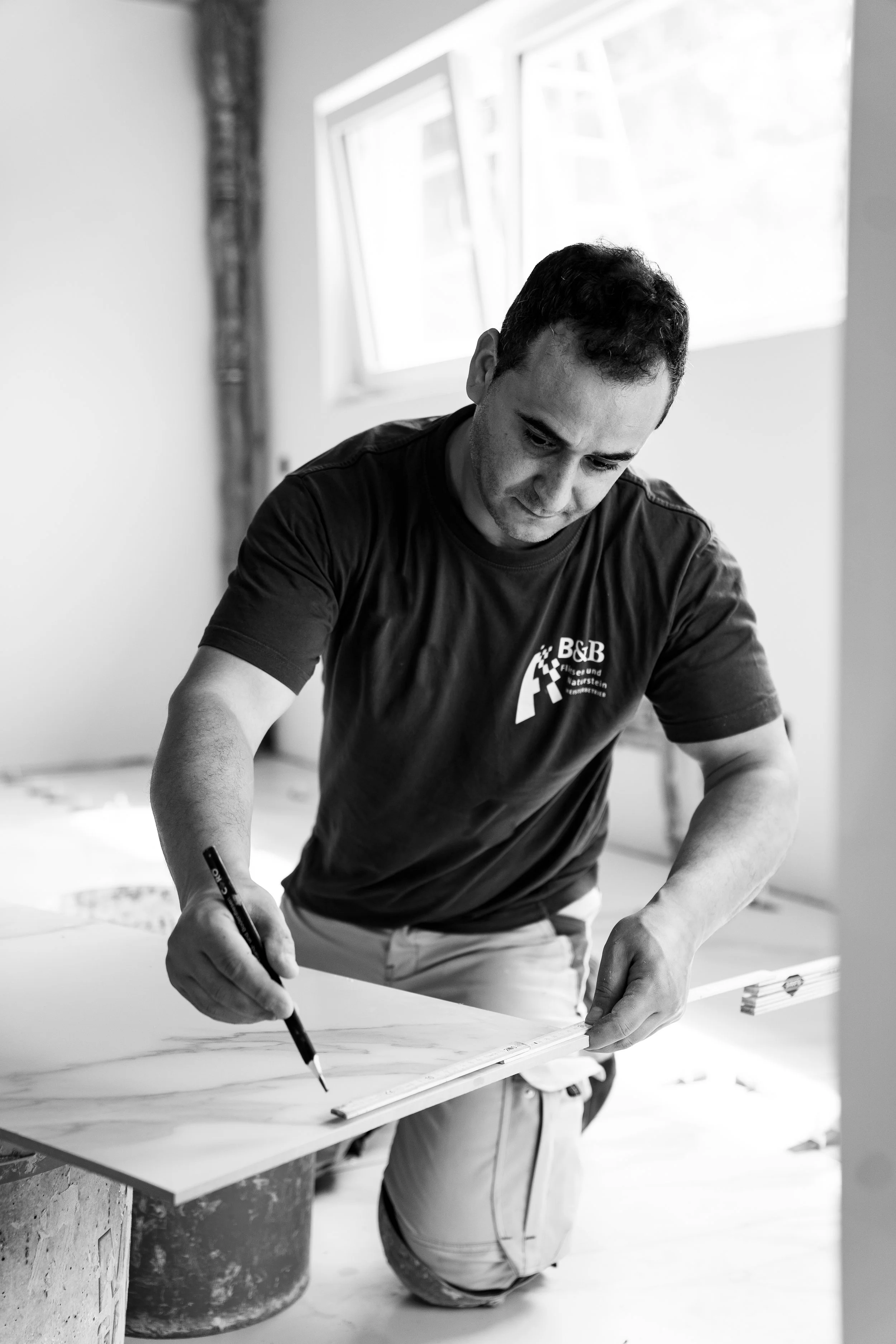 Ein Mann arbeitet an einem Renovierungsprojekt, er zeichnet auf eine Wand oder eine Platte, vermutlich bei Maler- oder Renovierungsarbeiten. Er trägt ein T-Shirt mit dem Logo "B&B".