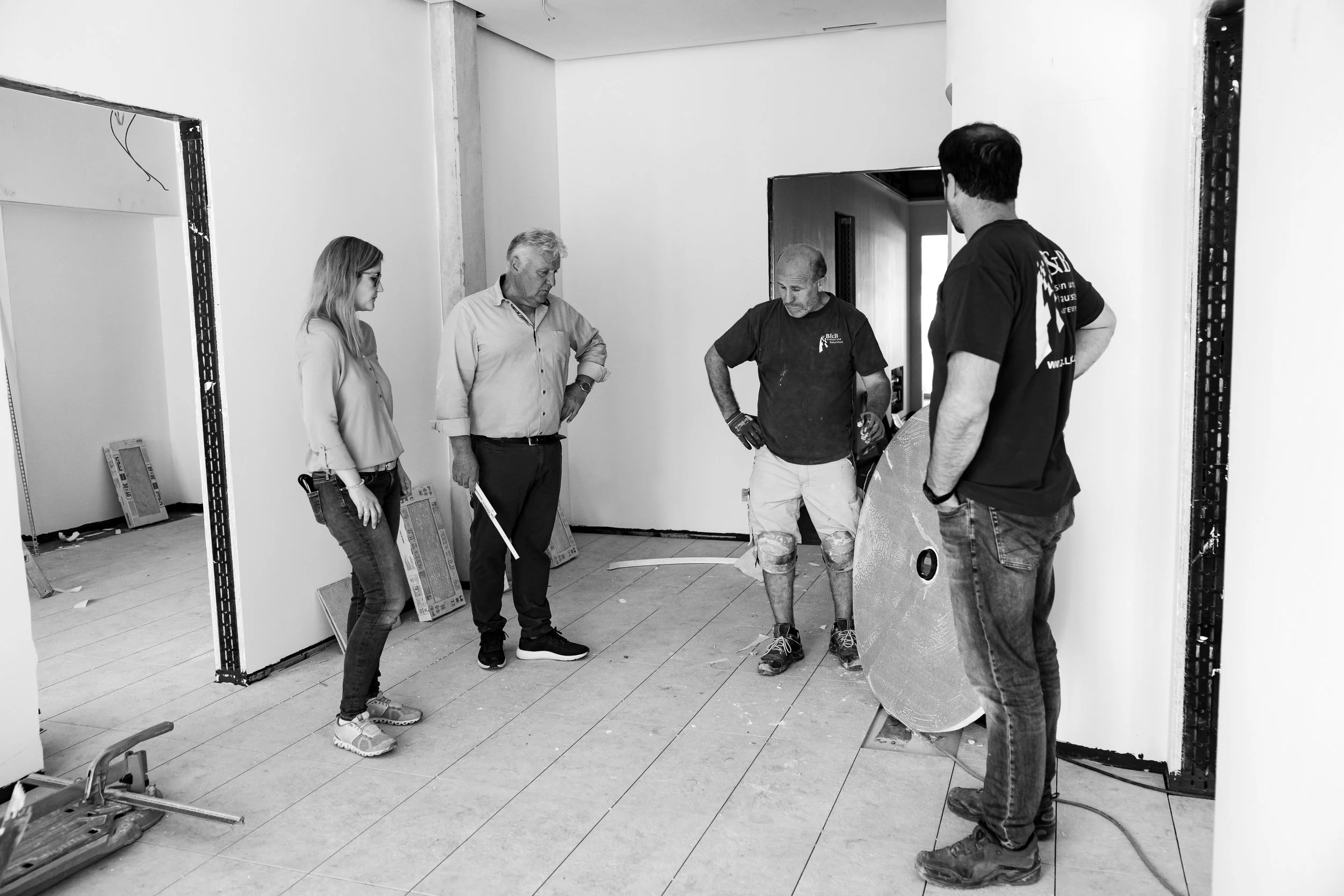 Fünf Menschen auf einer Baustelle während einer Besprechung, einige mit Werkzeugen, im Raum sind noch Baumaterialien sichtbar.