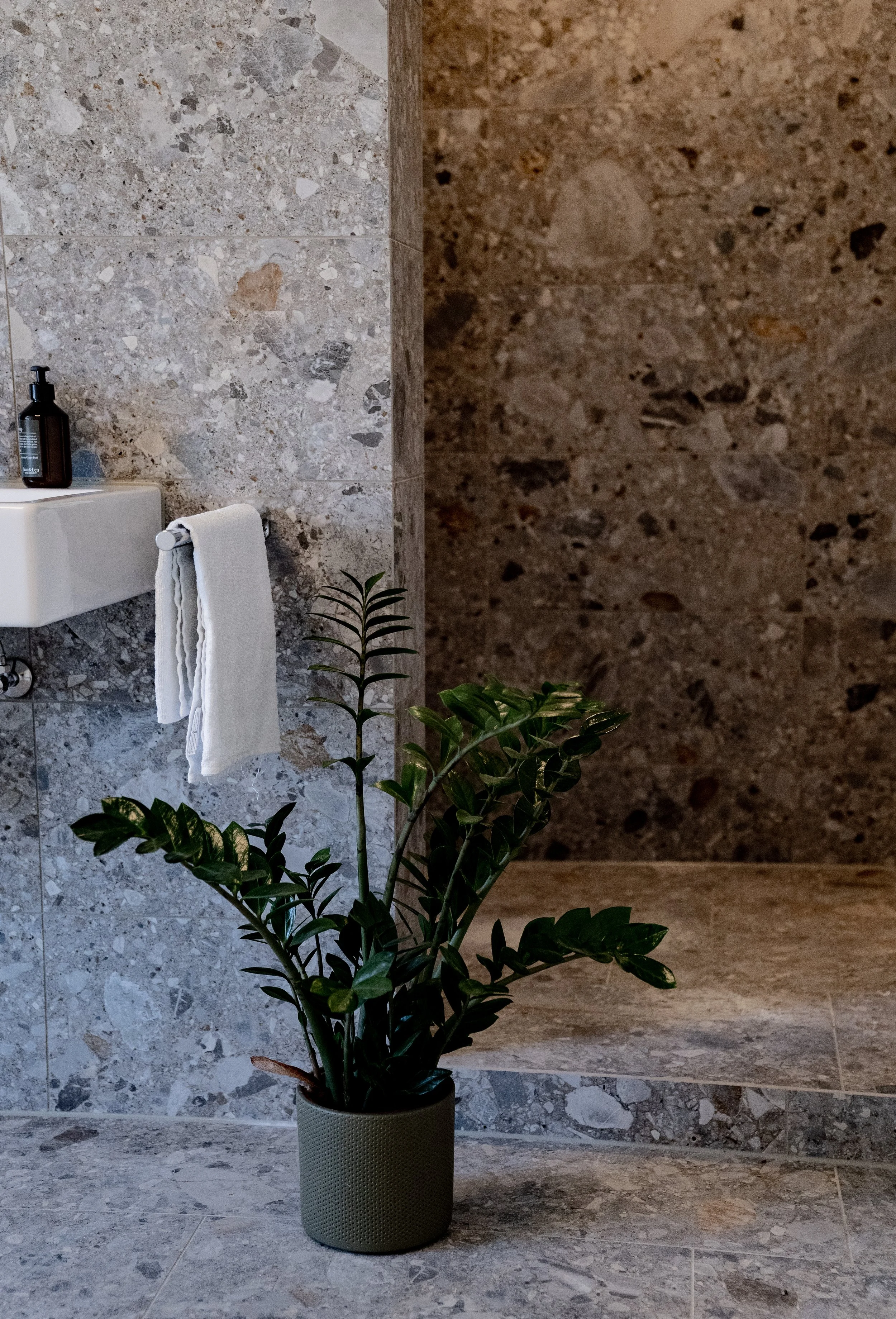 Ein Badezimmer mit einer Pflanze in einem großen Topf, einem Waschbecken mit einer schwarzen Seifenspender, einem weißen Handtuch, und Wänden und Boden aus grauem, gemustertem Marmor.