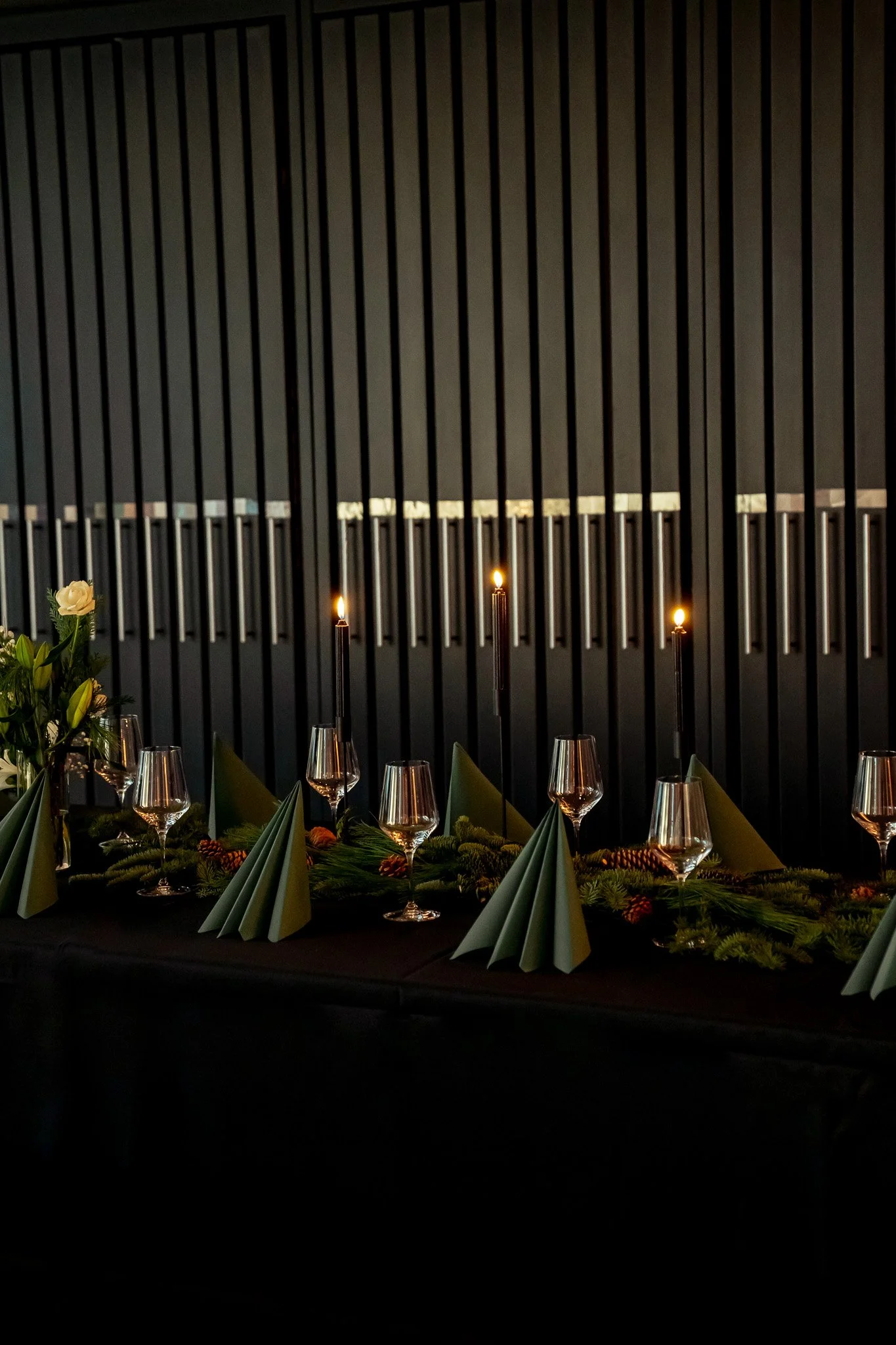 Ein festlich gedeckter Tisch mit Wein-, Wasser- und Sektgläsern, grünen Papierservietten, Kerzen und Weihnachtsdekoration auf einer schwarzen Tischdecke, vor einer dunklen vertikalen Holzverkleidung.