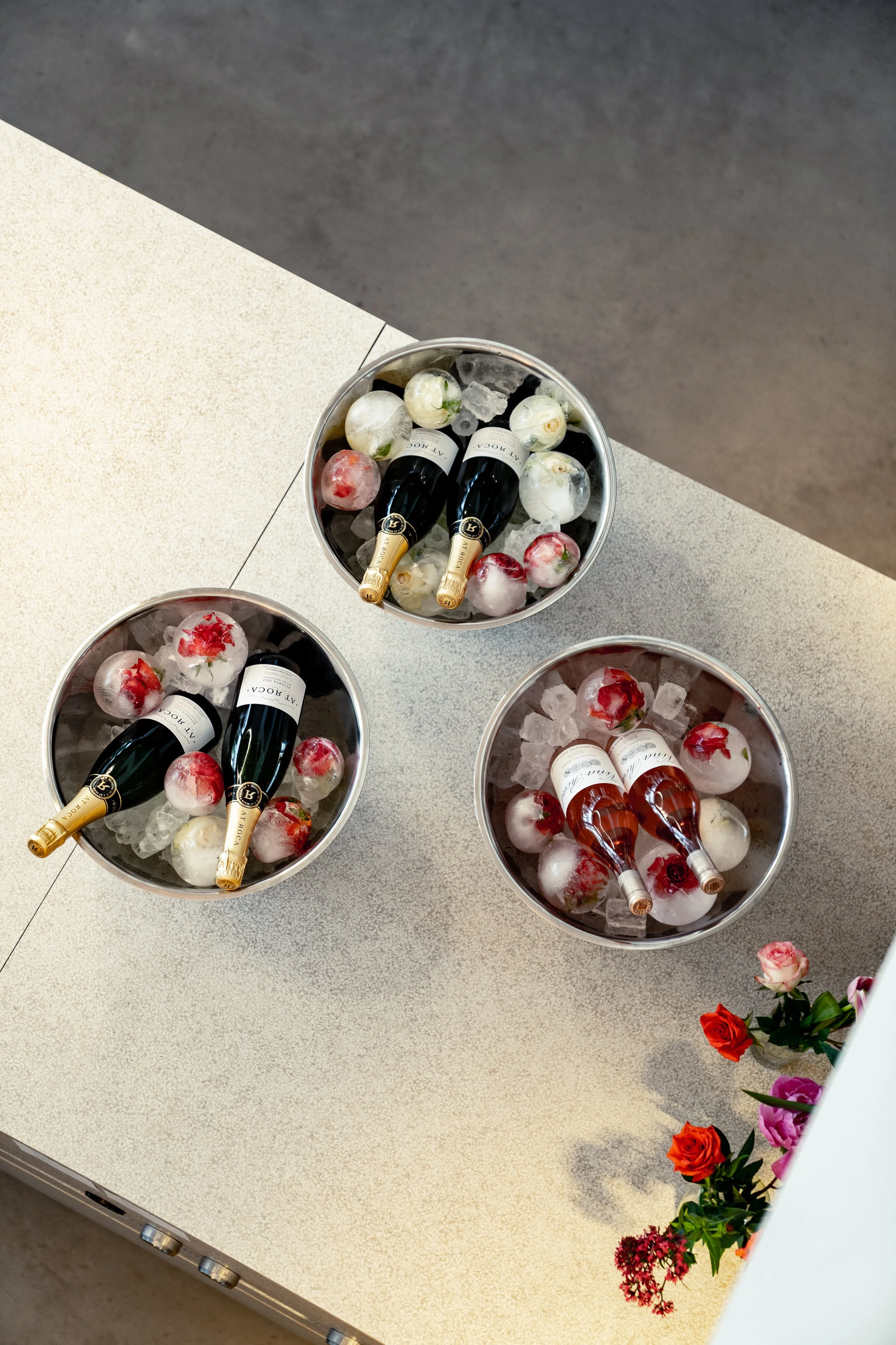 Drei Eisschalen mit Flaschen, Eiswürfeln und gefrorenen Rosen auf einem Tisch, dekoriert mit frischen Blumen.