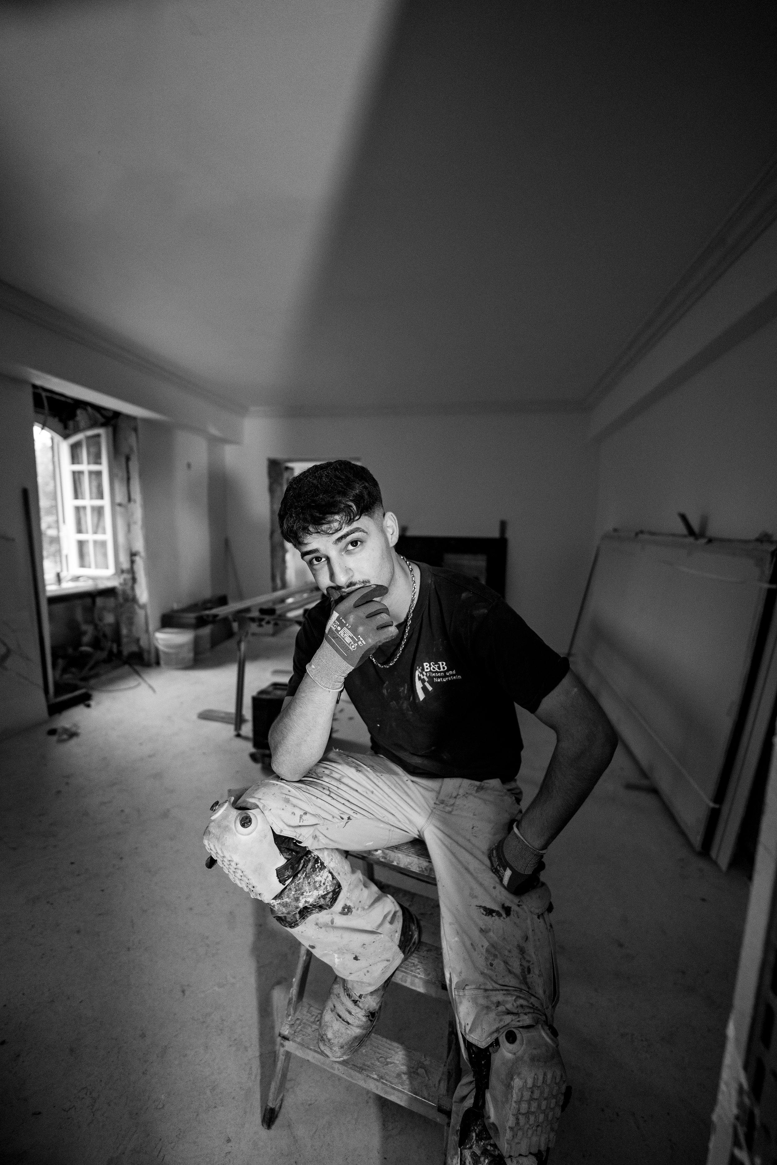 Junger Mann sitzt auf einer Holzleiter in einem Raum mit Baumaterialien, trägt Arbeitshosen und Handschuhe, schaut in die Kamera, schwarz-weiß, Baustellenumgebung.