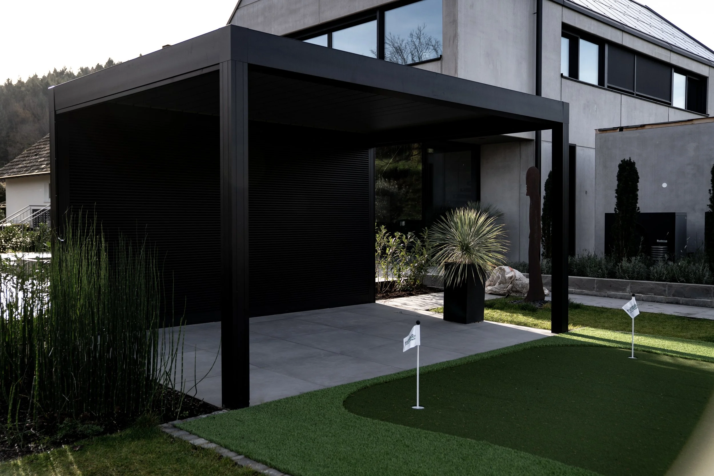 Moderne Hausvorderseite mit Carport, grünem Rasen, kleineren Bäumen und Sträuchern, bei bewölktem Himmel.