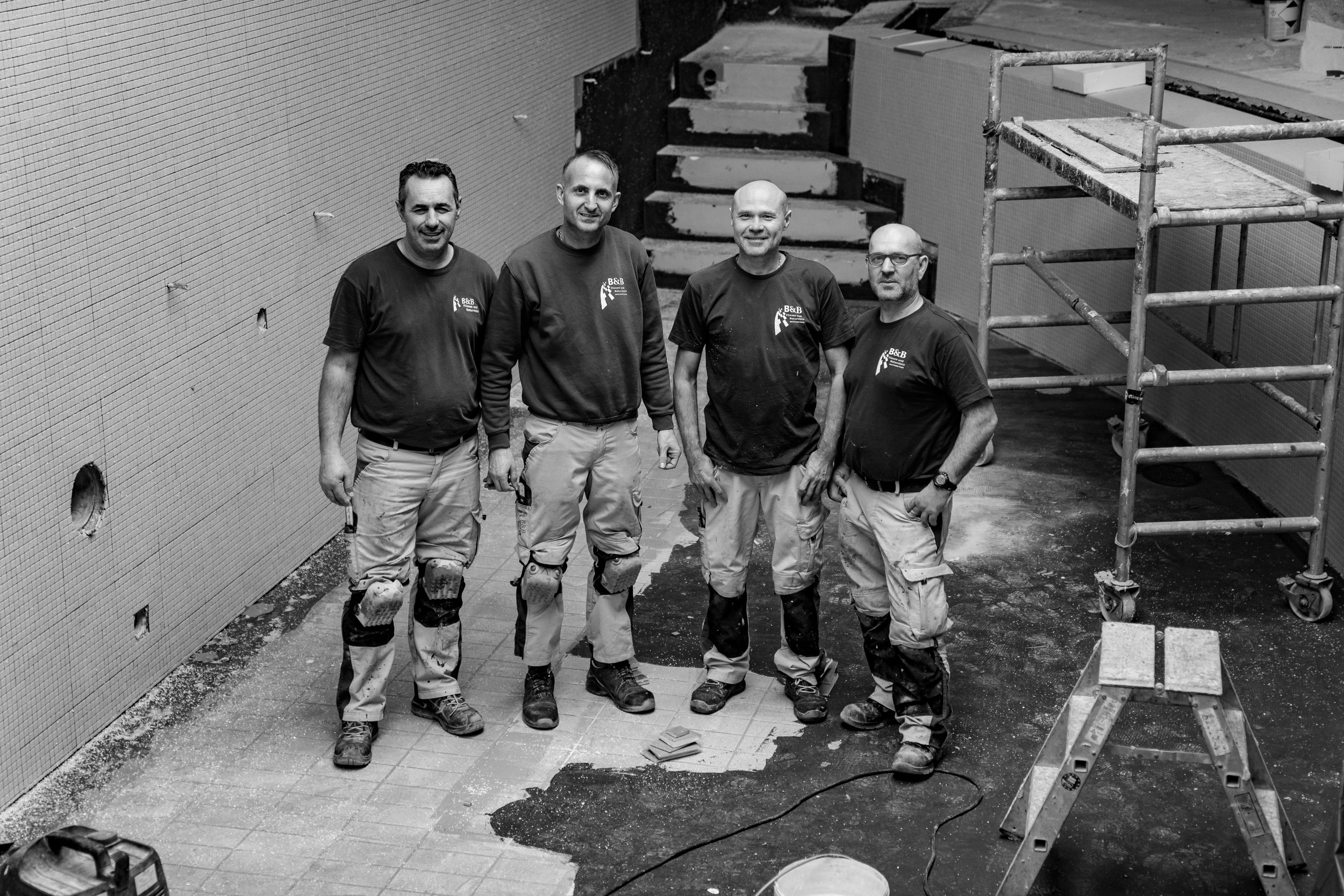 Vier Bauarbeiter stehen in einem Raum, der renoviert wird, vor einer Wand mit Fliesen, einige Werkzeuge und eine gerüste. Es handelt sich um eine Schwarz-Weiß-Fotografie.