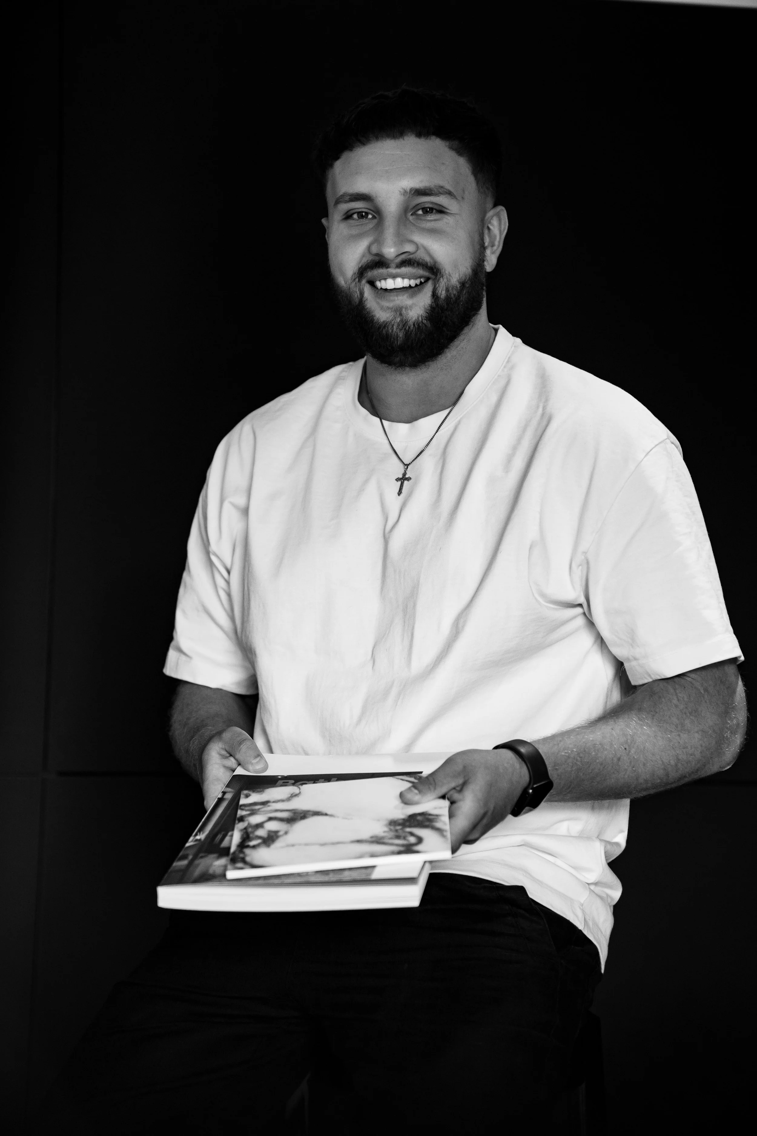 Junger Mann mit Bart und Kreuzkette sitzt vor dunklem Hintergrund, hält Bücher in den Händen, trägt ein weißes T-Shirt und ein Armband. Er lächelt in die Kamera.