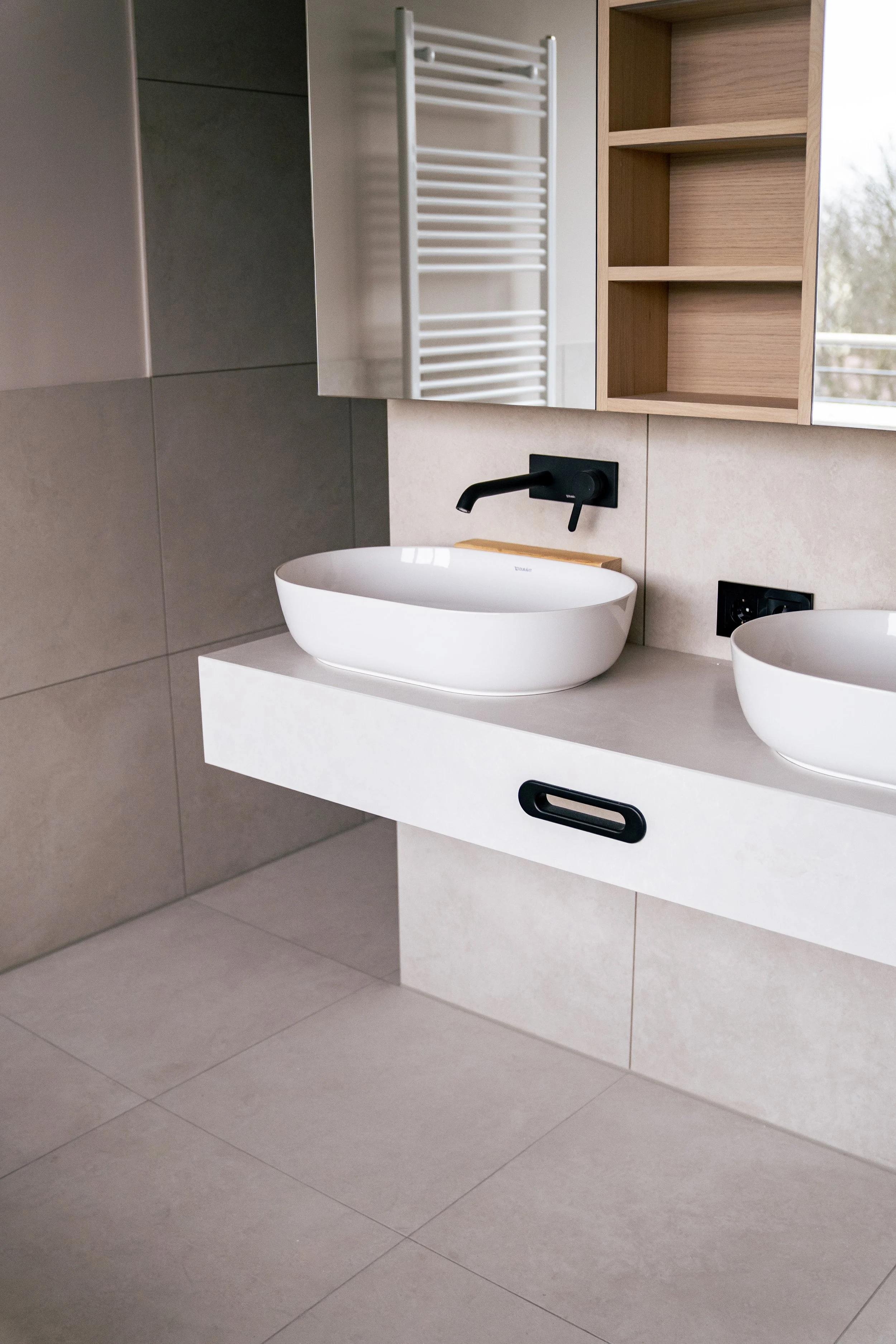 Moderner Badezimmerbereich mit doppeltem Waschtisch, weißen ovalen Waschbecken, schwarzen Armaturen, angeschlossenem schwarzen Stromanschluss, offener Holzregal und einem beheizten Handtuchhalter an der Wand.