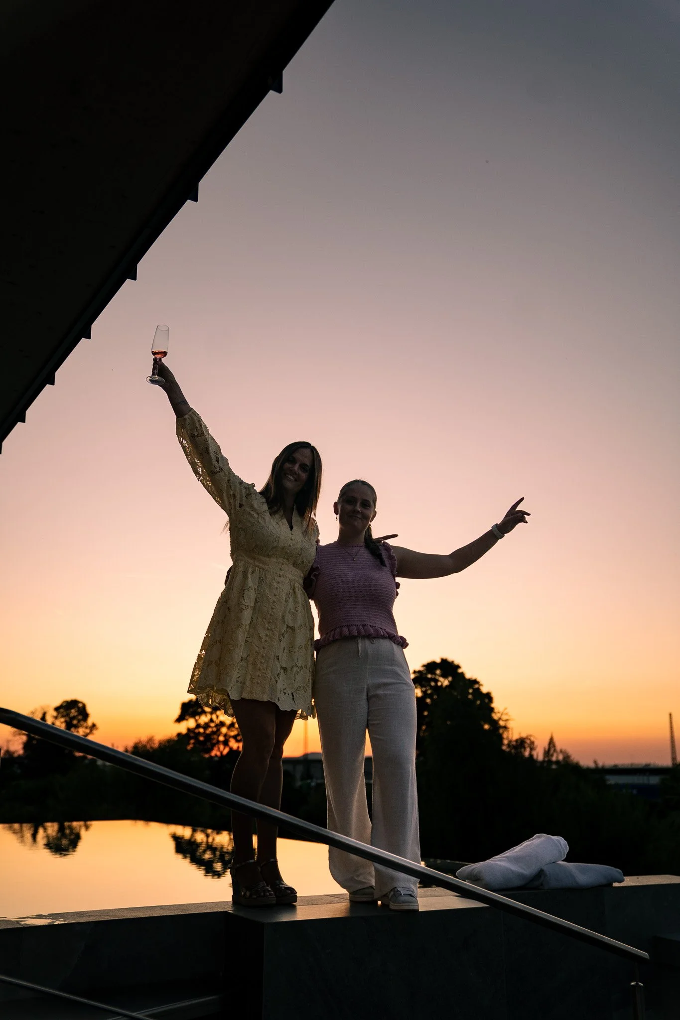 Zwei Frauen stehen bei Sonnenuntergang auf einer Plattform am Wasser, eine hält ein Glas Wein hoch.