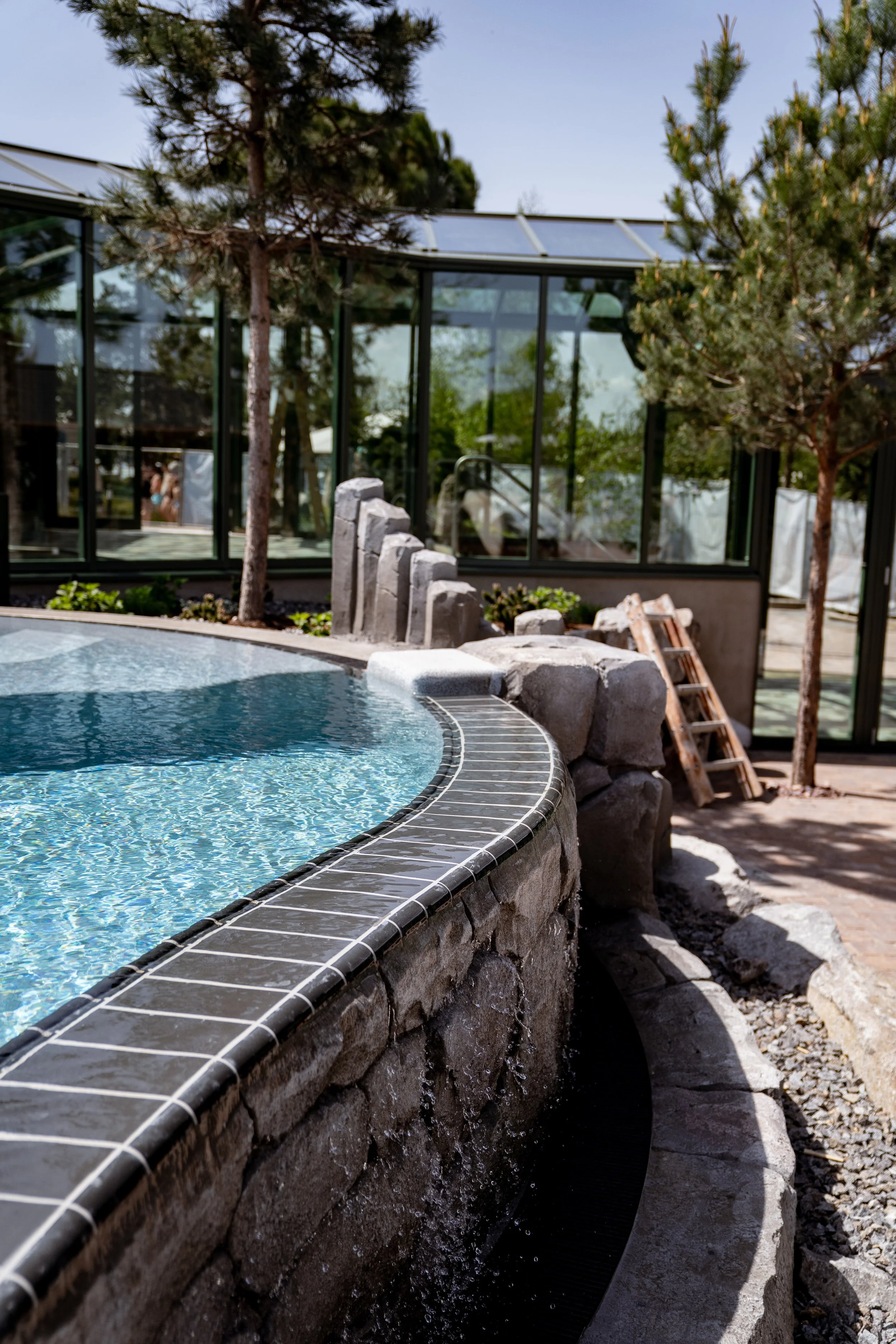 Ein Whirlpool im Freien mit Wasser, umgeben von Steinen und kleinen Bäumen, vor einem modernen Glashaus bei sonnigem Wetter.