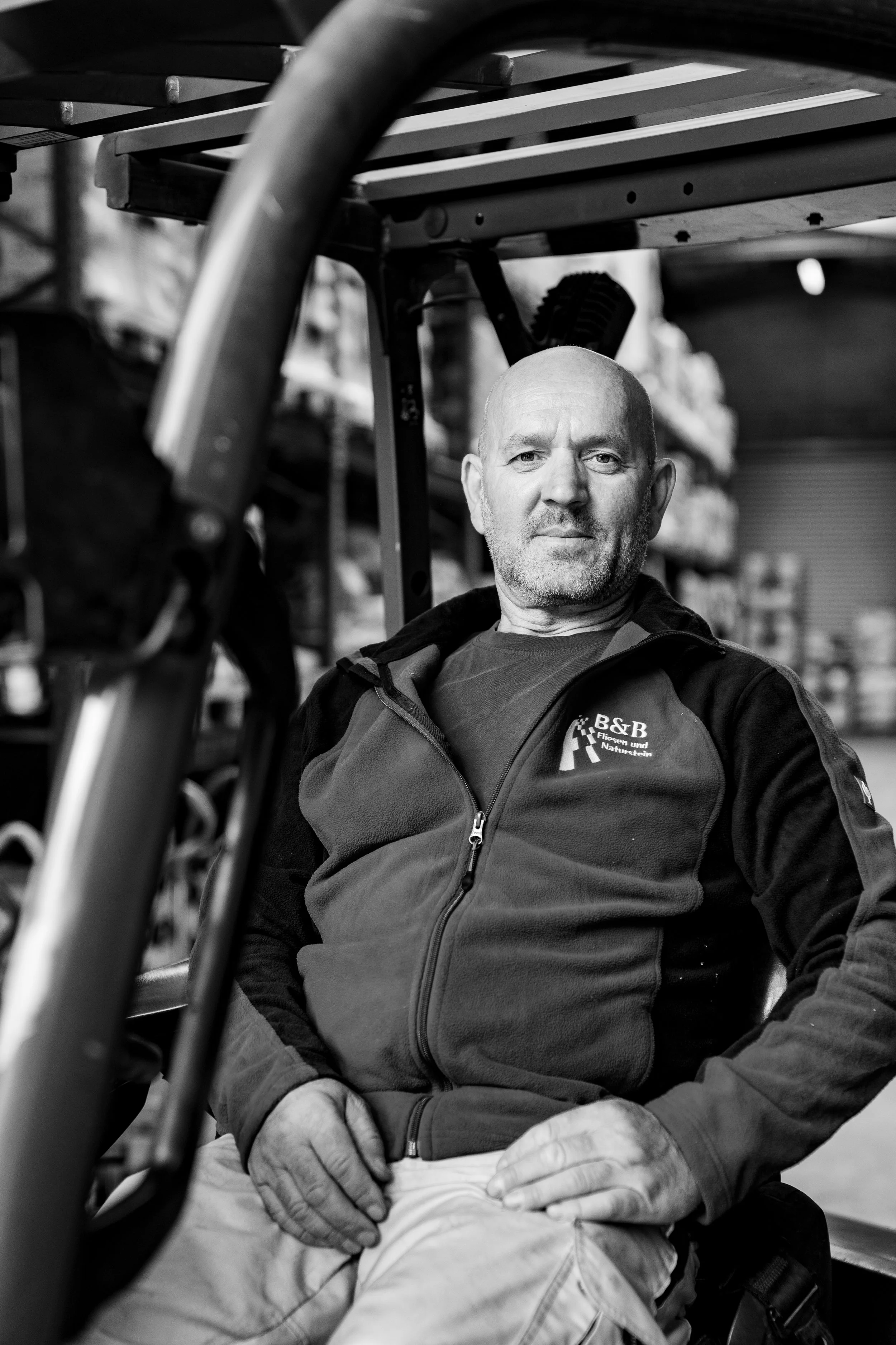 Ein Mann sitzt in einem Lagerraum oder Werkstatt, trägt eine hoodie mit einem Logo und schaut in die Kamera.