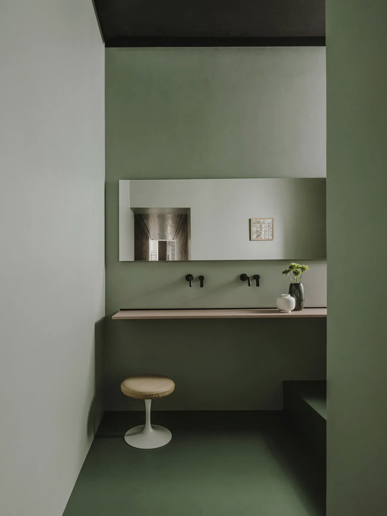 Ein minimalistisches Badezimmer mit grünen Wänden, einem großen rechteckigen Spiegel, einem Waschtisch mit zwei Wasserhähnen, einer Vase mit Blumen und einem kleinen weißen Behälter auf dem Waschtisch, einem Stuhl und einer kleinen Sitzbank.