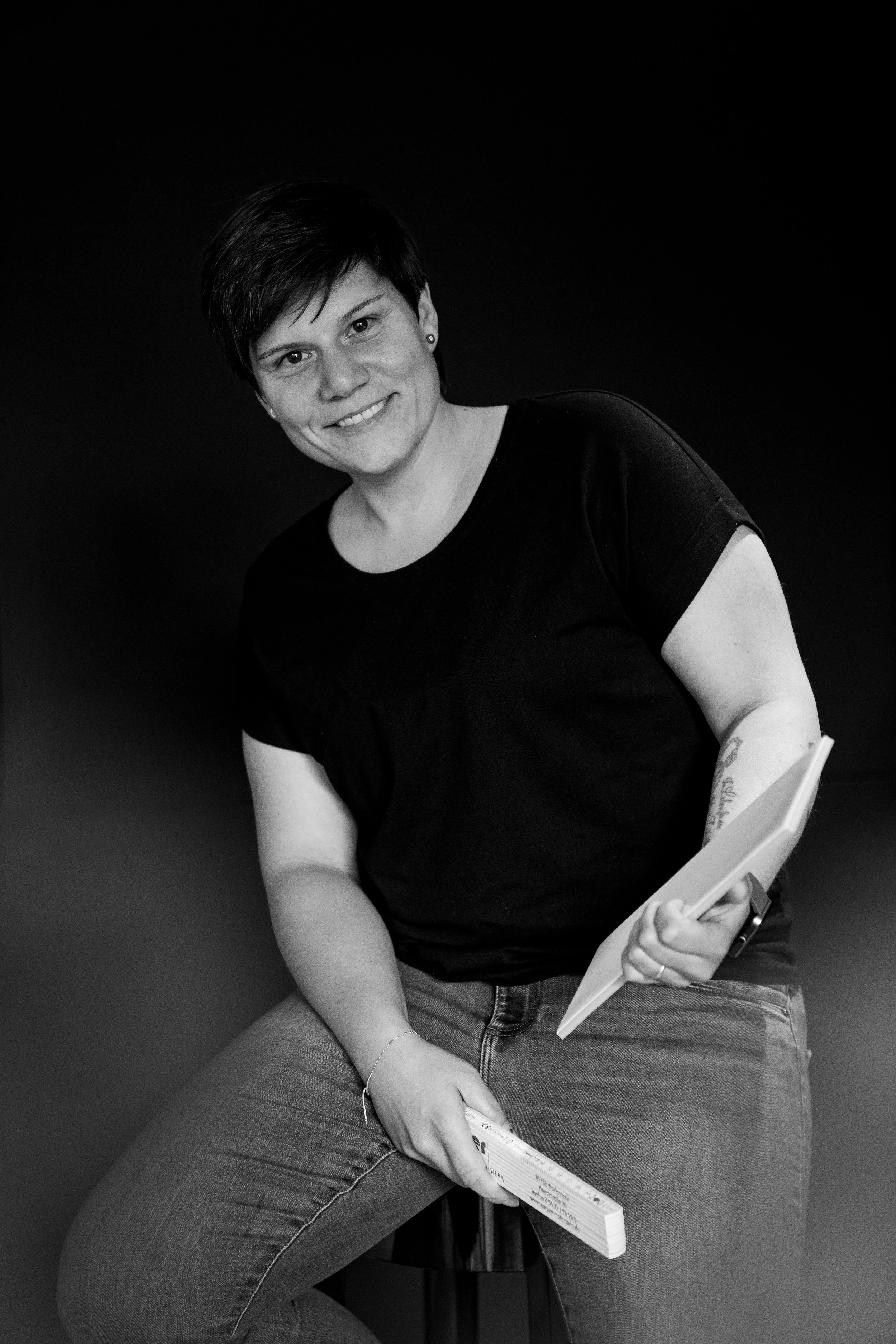 Schwarz-weiß Portrait einer lächelnden Frau mit kurzen schwarzen Haaren, sitzend auf einem Hocker, trägt ein schwarzes T-Shirt und Jeans, hält ein Notizbuch und eine Zeitschrift in den Händen, gegen einen dunklen Hintergrund.