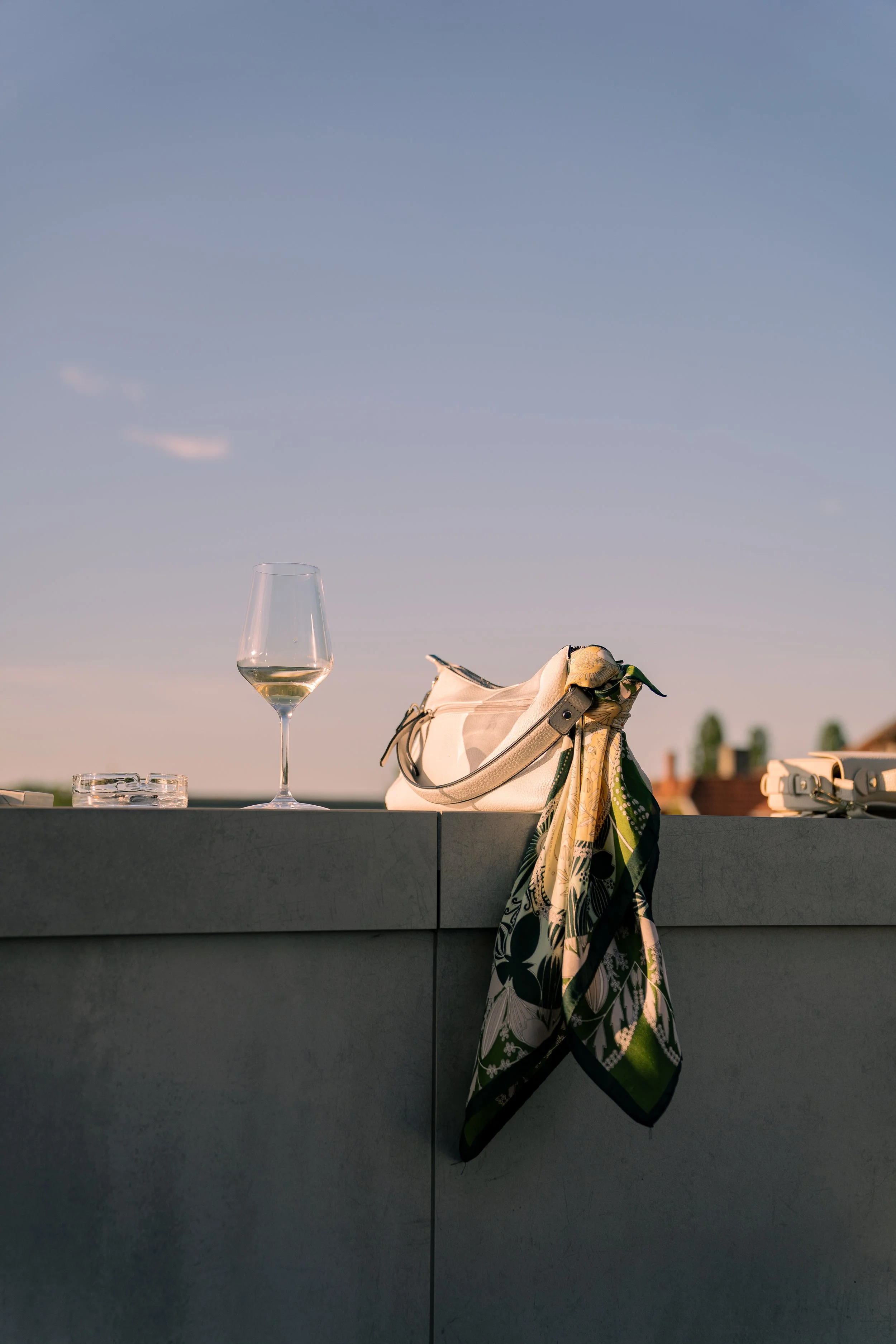 Auf einem Balkon steht ein Weinglas mit weißem Wein, eine Tasche mit einem Schal hängt an der Mauer, im Hintergrund sind Dächer und Bäume bei Sonnenuntergang sichtbar.