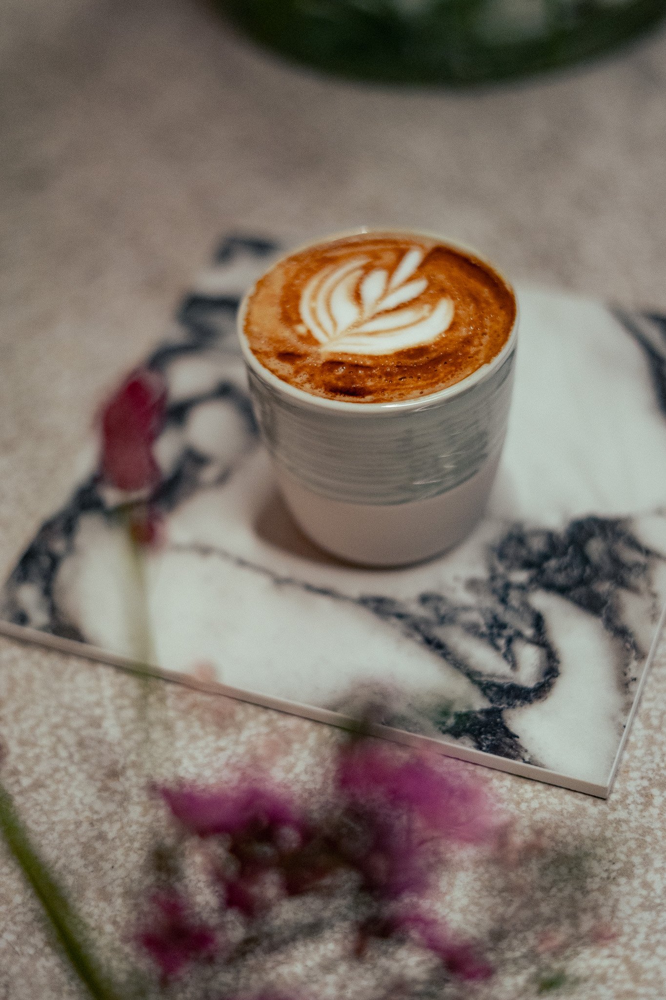 Ein Cappuccino mit Milchschaumdekoration in einer weißen Tasse auf einem Marmorboden. Im Hintergrund sind zwei pinkfarbene Blumen leicht sichtbar.