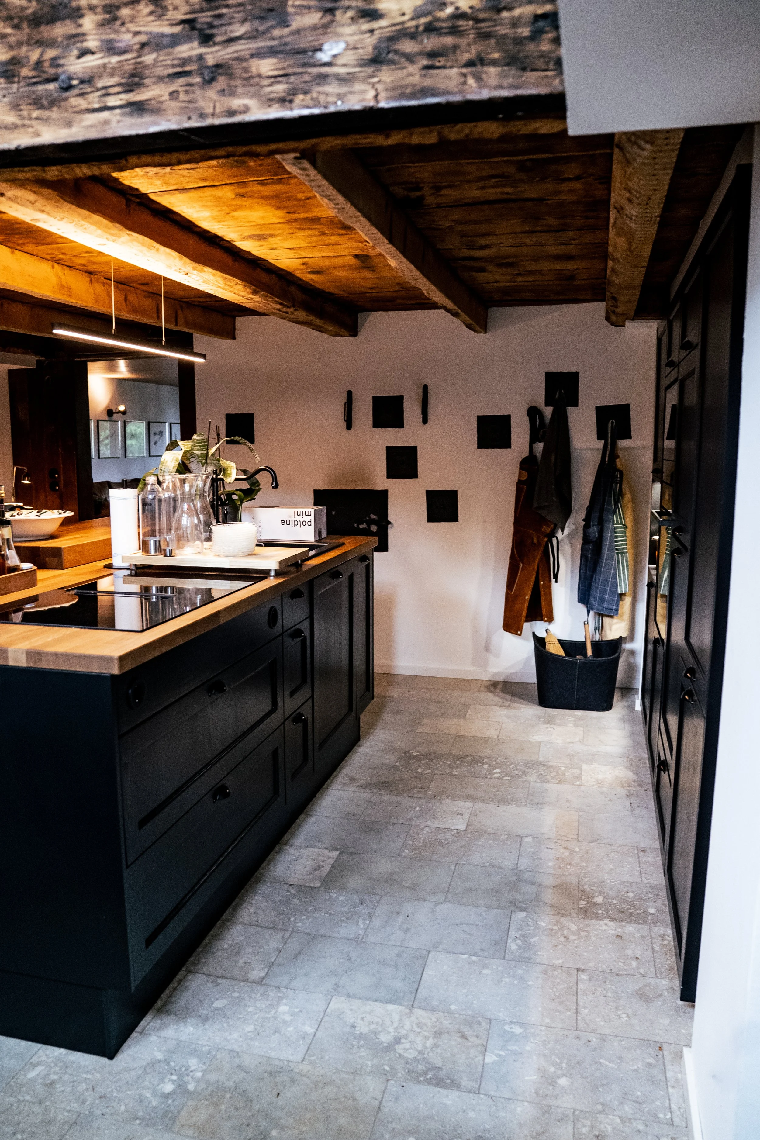 Moderner Küchenbereich mit schwarzen Schränken, Holzarbeitsplatte, Holzdecke und einer weißen Wand mit schwarzen Dekorationen, inklusive Wandhaken mit Küchenschürzen