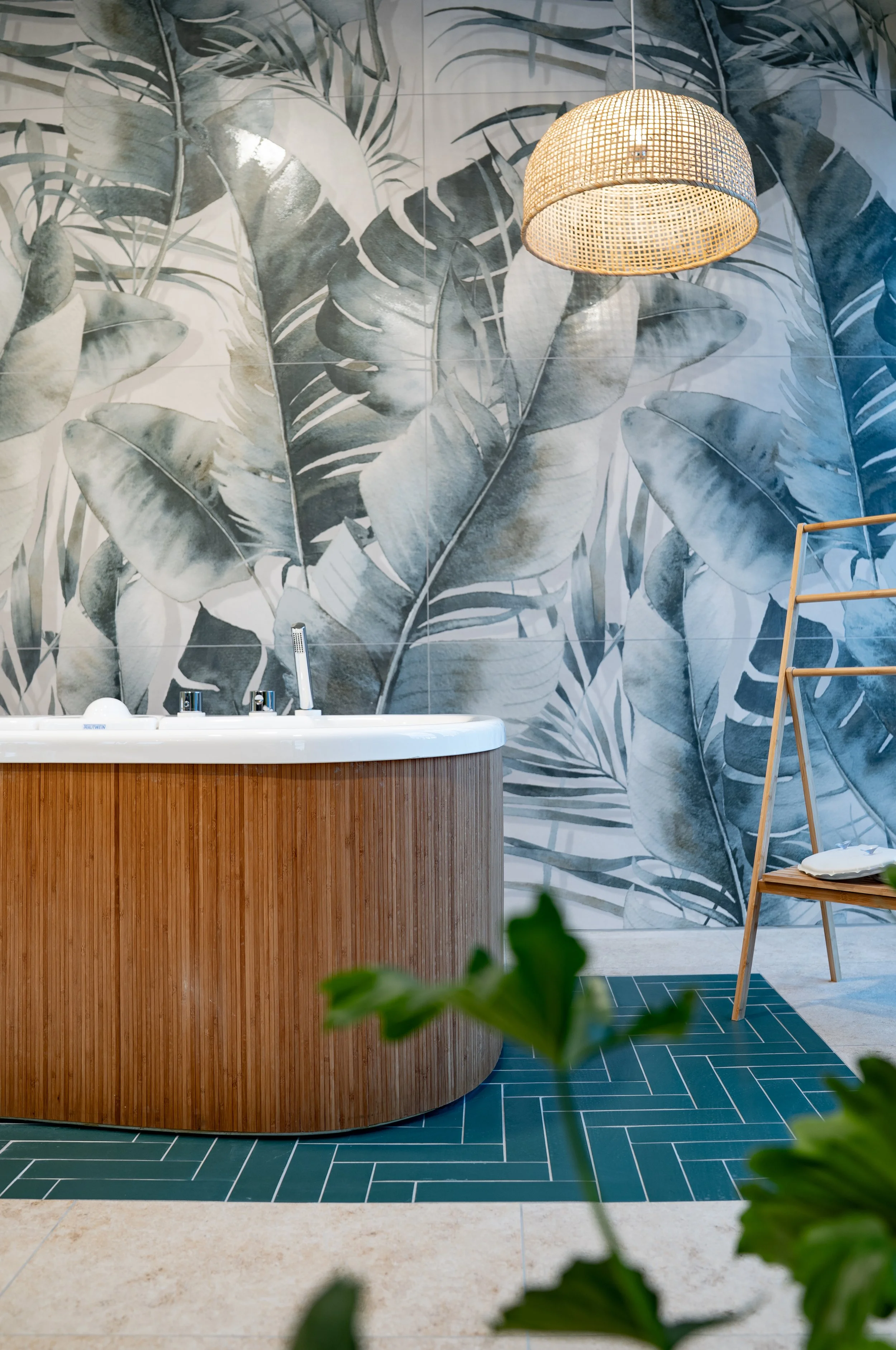 Ein Badezimmer mit einer großen, runden Wanne aus Holz, einer Wandtapete mit großflächigem Monstera-Blätter-Motiv, einer hölzernen Stehlampe, einem Holzstuhl mit weißen Handtüchern, grünen Pflanzen im Vordergrund und blauen Wandfliesen auf dem Boden.