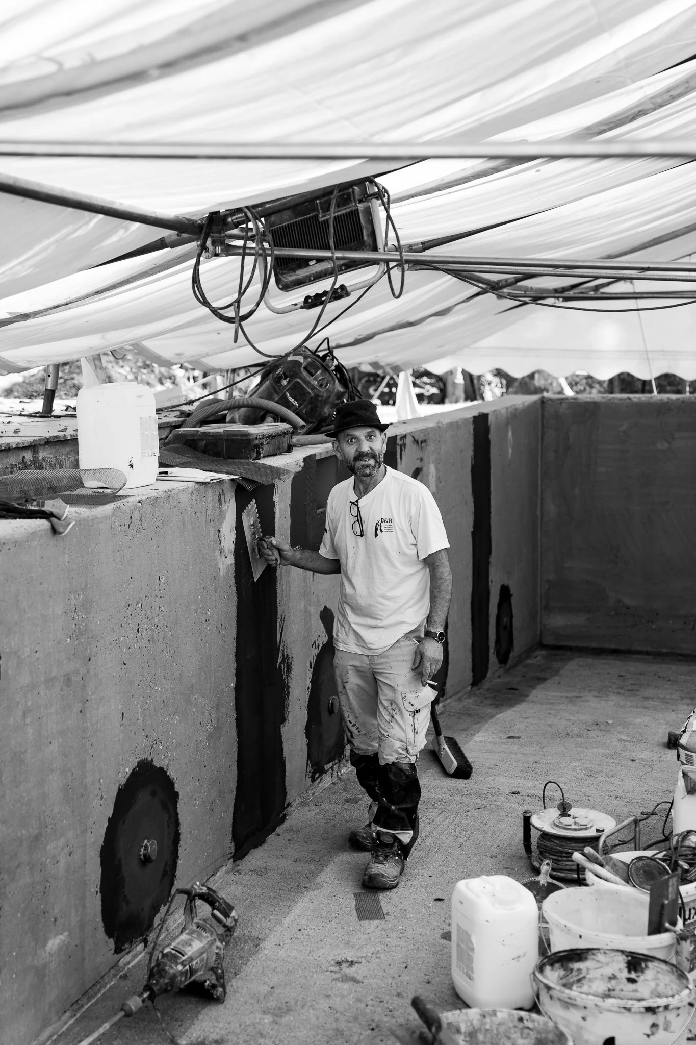 Ein Mann mit Bart und Brille hängt an einer Mauer und arbeitet an einer Baustelle. Er trägt ein weißes T-Shirt, Arbeitshosen und einen Hut.