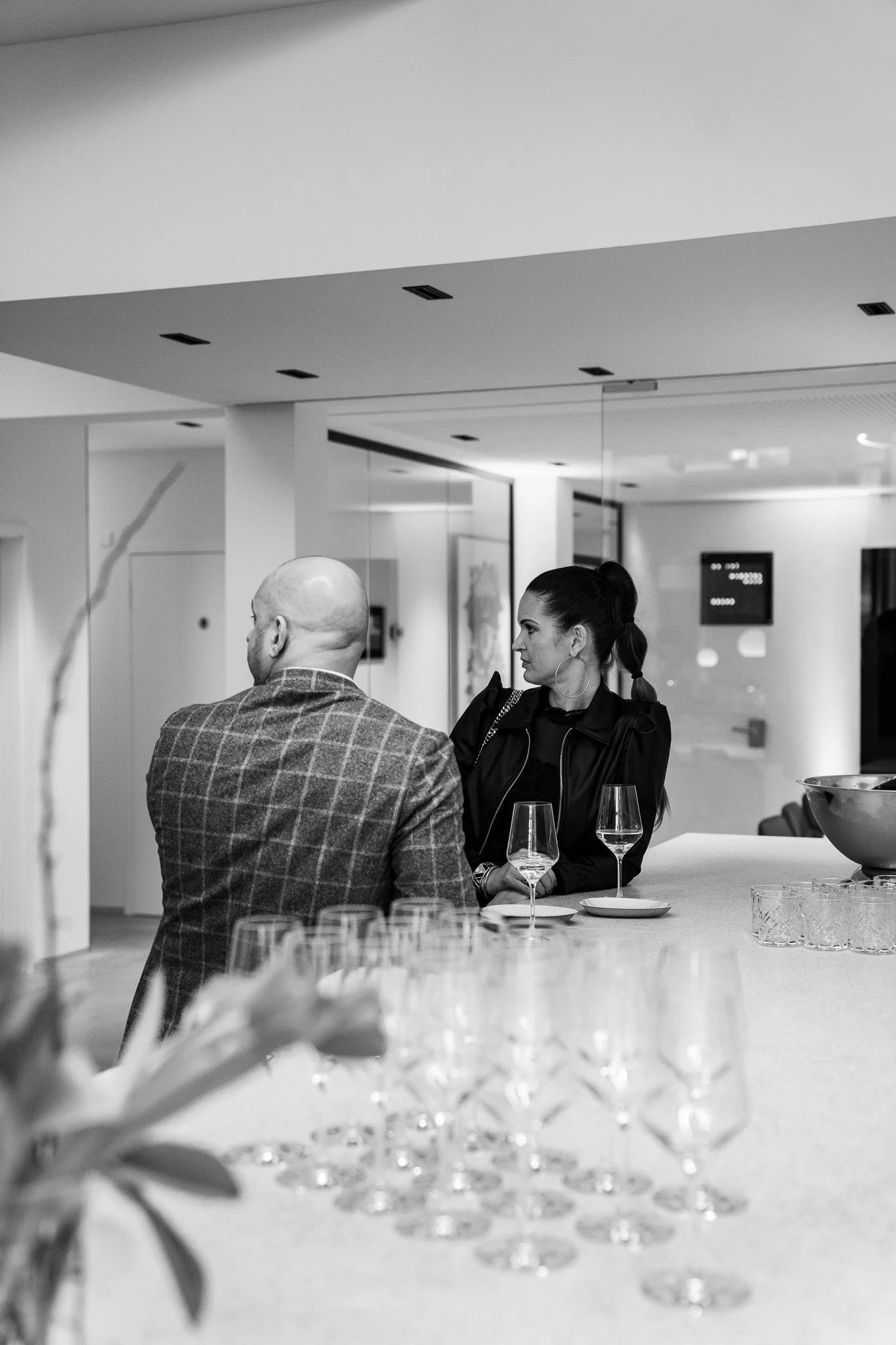 Zwei Personen, ein Mann mit Glatze und kariertem Anzug und eine Frau mit dunklen Haaren, sitzen an einer Bar mit Gläsern, im Hintergrund eine moderne, helle Lokalumgebung.