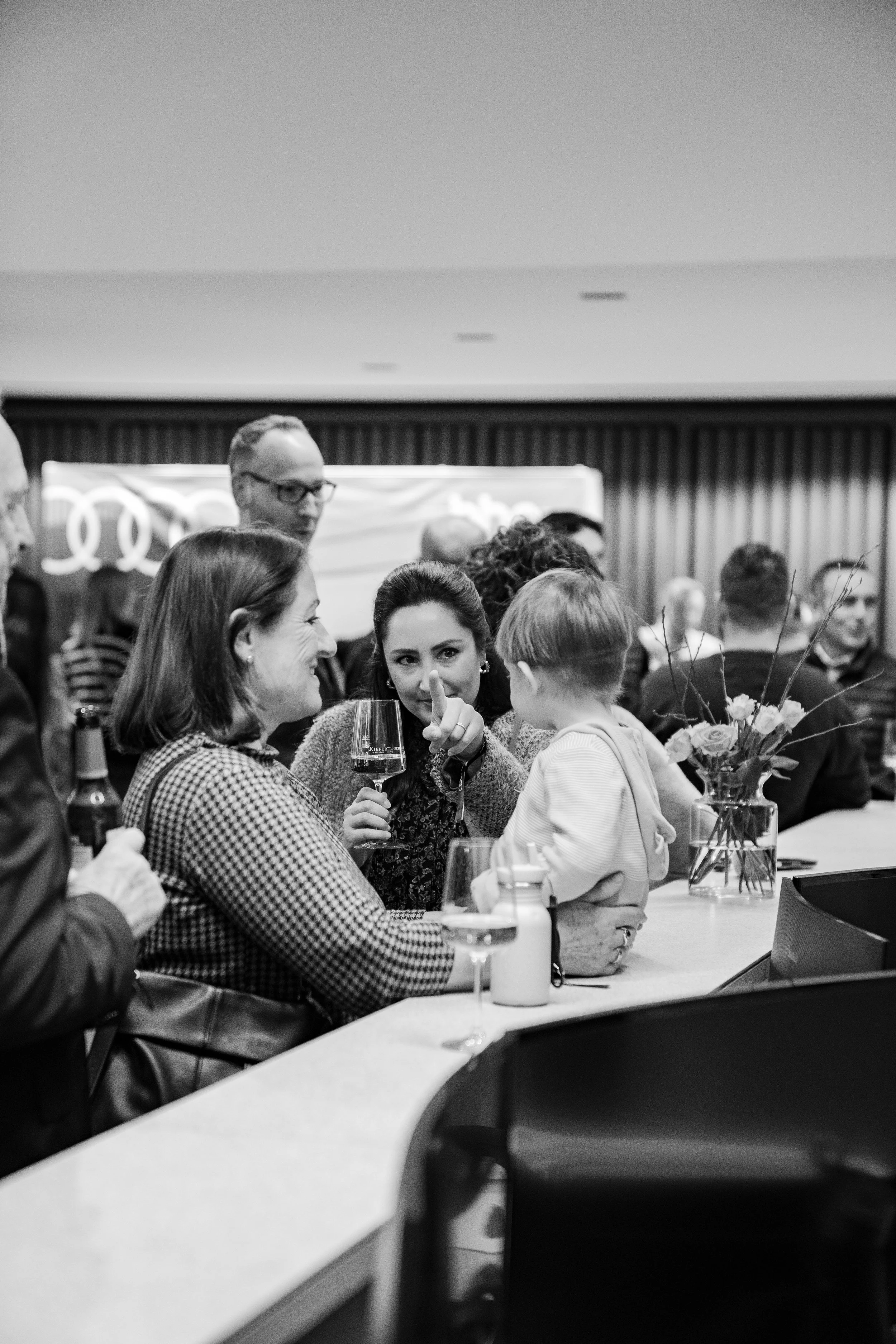Menschen sitzen an einem Tresen, eine Frau zeigt mit dem Finger auf eine kleine Frau mit einem Glas Wein in der Hand. Es ist ein geselliger Raum mit weiteren Personen im Hintergrund und einem Blumenstrauß auf dem Tisch.